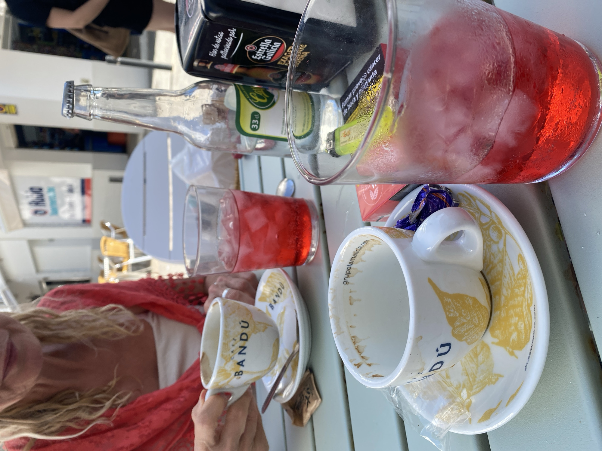 A close-up shot captures two cups of coffee and two red drinks with ice on a light green table, suggesting an outdoor cafe setting in Conil de la Frontera, Spain. In the foreground, a white coffee cup with yellow leaf-like designs and the text "BANDU" sits on a matching saucer. A small spoon rests on the saucer, and a sugar packet is partially visible. To the right, a red drink in a frosted glass is filled with ice. Behind these, another white cup, presumably also with coffee remnants, is partially visible, and a second red drink in a glass is to its right.
A clear glass bottle with a metallic cap, labeled with "33 cl" and "Cerveza," stands between the cups. To its right is a black napkin dispenser with the "Estrella Galicia" logo, indicating a possible connection to a brewery or bar. In the background, a woman wearing a red shawl over a white top is visible, her hands holding one of the coffee cups. The setting appears to be a street-side cafe, with blurred white buildings and tables and chairs in the distance, consistent with the architecture and atmosphere of Conil de la Frontera. The lighting suggests daytime.