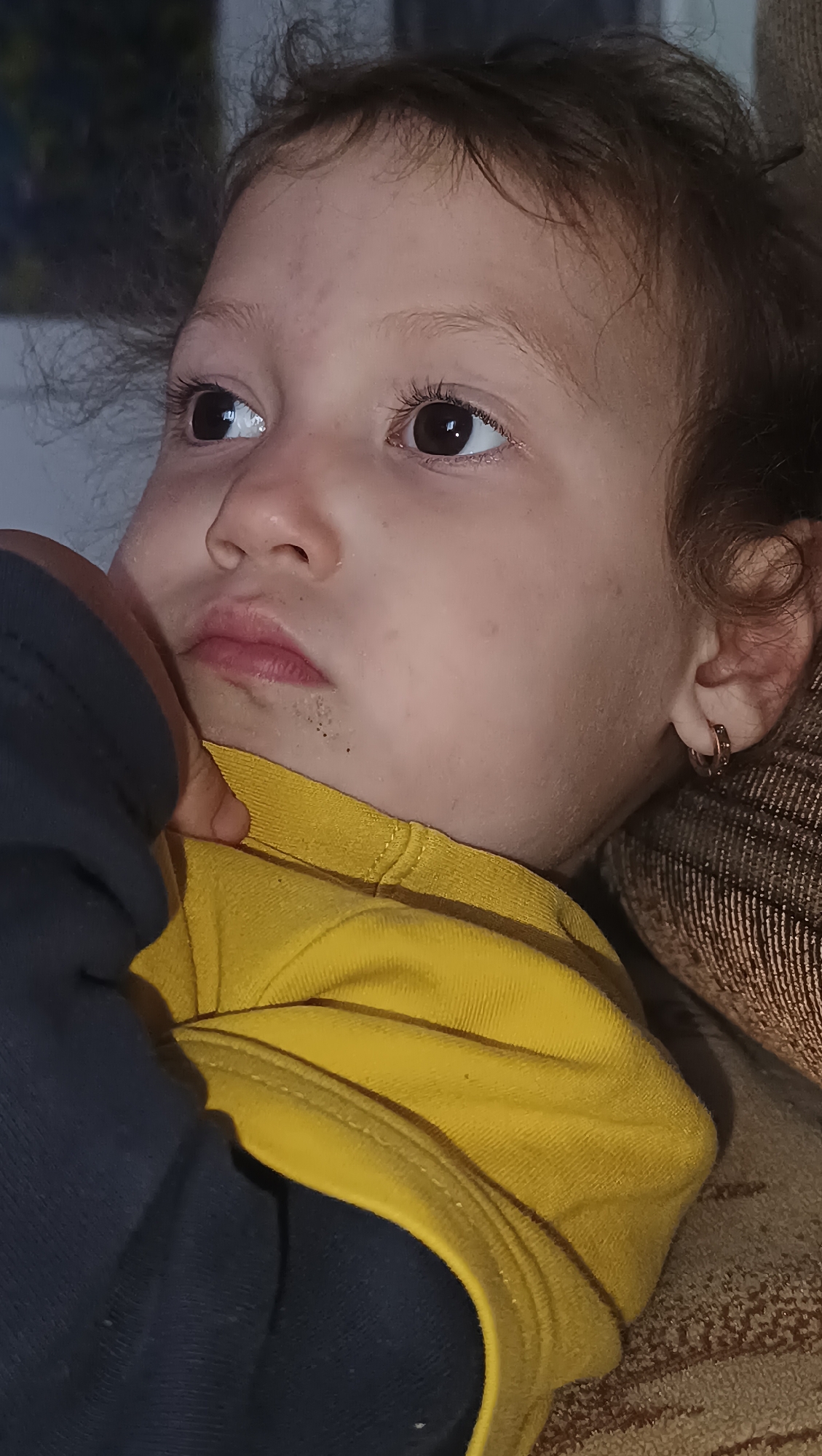 The image is a close-up portrait of a young child with dark, curly hair and large, dark eyes. The child is wearing a yellow shirt with a dark blue sleeve visible on the left. The child's left hand is resting on their chin and chest. There are a few dark spots on the child's chin. The child is wearing a small hoop earring in their right ear. The background is blurred, showing a textured surface that appears to be a couch or cushion. The lighting is dim, with reflections visible in the child's eyes. The image does not provide any information or context about the city of Karsha, Russia.