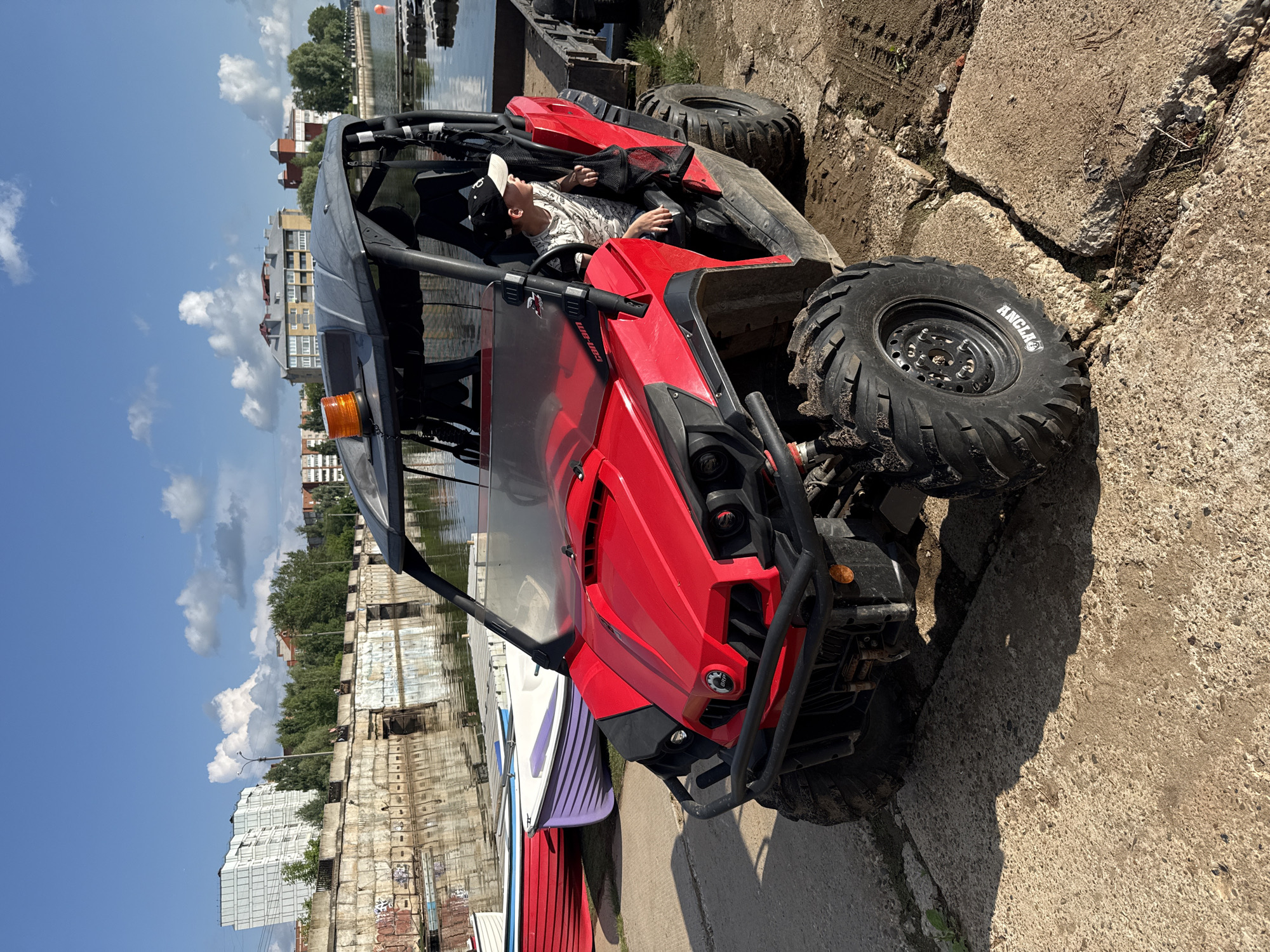 A young boy wearing a black and white baseball cap and a patterned t-shirt sits in the driver's seat of a bright red Can-Am all-terrain vehicle (ATV). The ATV is parked on a concrete surface next to a body of water, likely a river or canal. Several small boats, colored red, white, and purple, are lined up near the water's edge. In the background, a concrete embankment with graffiti lines the waterfront, and beyond that, buildings and trees are visible under a partly cloudy blue sky. The scene appears to be set in an urban area along a waterway.