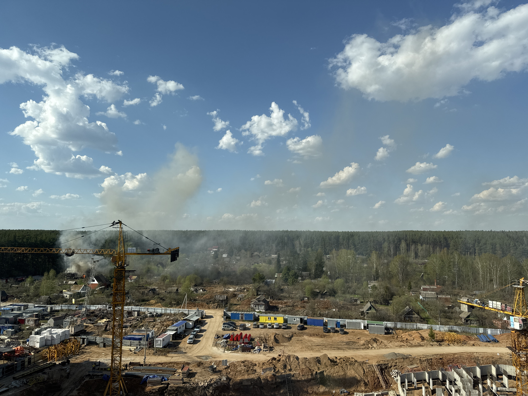 The image shows a construction site in Izhevsk, Russia, under a blue sky with scattered white clouds. Two yellow cranes stand prominently on the left and right sides of the frame. In the center, smoke rises in the distance from what appears to be a fire in a residential area with scattered houses. The construction site has dirt ground, exposed concrete structures, and various temporary buildings, containers, and construction materials. There are also several cars parked nearby. The background features a dense forest.