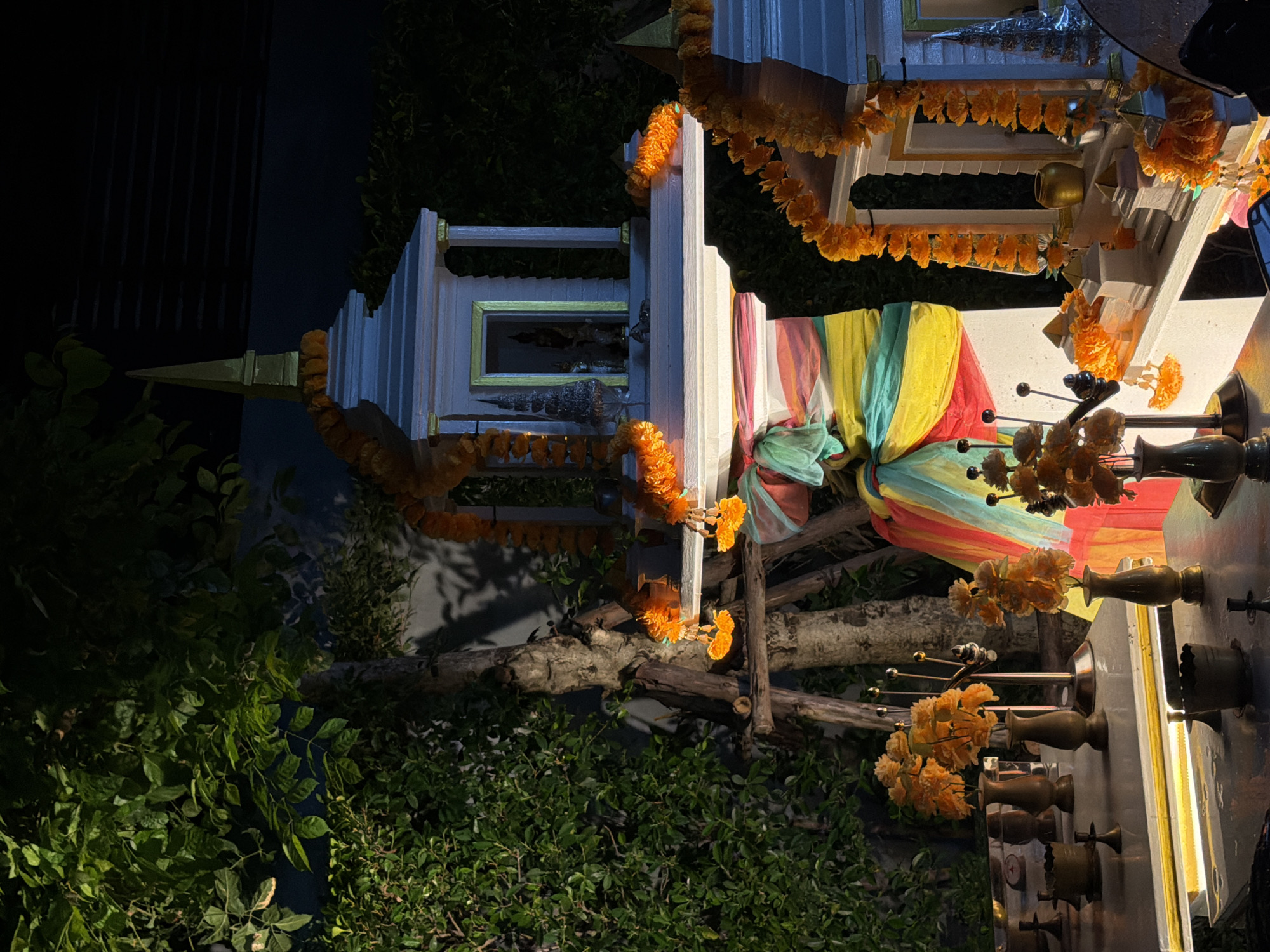 This nighttime image captures a traditional Thai spirit house (san phra phum) within a lush, green outdoor setting, likely a garden or property in Bangkok, Thailand. The scene is brightly illuminated by artificial light against the dark backdrop of night.
In the center-right of the frame stands a prominent white spirit house with a pointed golden spire and golden trim. It is richly decorated with numerous garlands of vibrant orange marigold flowers, draped across its roofline and structural elements. To its left, a smaller, similar spirit house is also adorned with marigold garlands. Inside this smaller structure, a seated figurine, likely a deity or guardian spirit, is visible.
A key visual element is a multi-colored fabric sash, consisting of horizontal stripes of red, yellow, green, and blue, tied around a central support or tree trunk located between the two spirit houses. This fabric also features some attached marigold flowers.
On a white platform in the foreground, directly in front of the main spirit house, several traditional offerings are arranged. These include various brass or golden vessels, some holding more marigold flowers, and others appearing to be ornate incense holders with thin, dark rods protruding from them.
The entire setup is nestled amidst dense, dark green foliage and tree branches that frame the scene from the bottom and extend into the background. The low light and dramatic illumination create an atmosphere of reverence and spiritual significance. There is no visible text in the image.