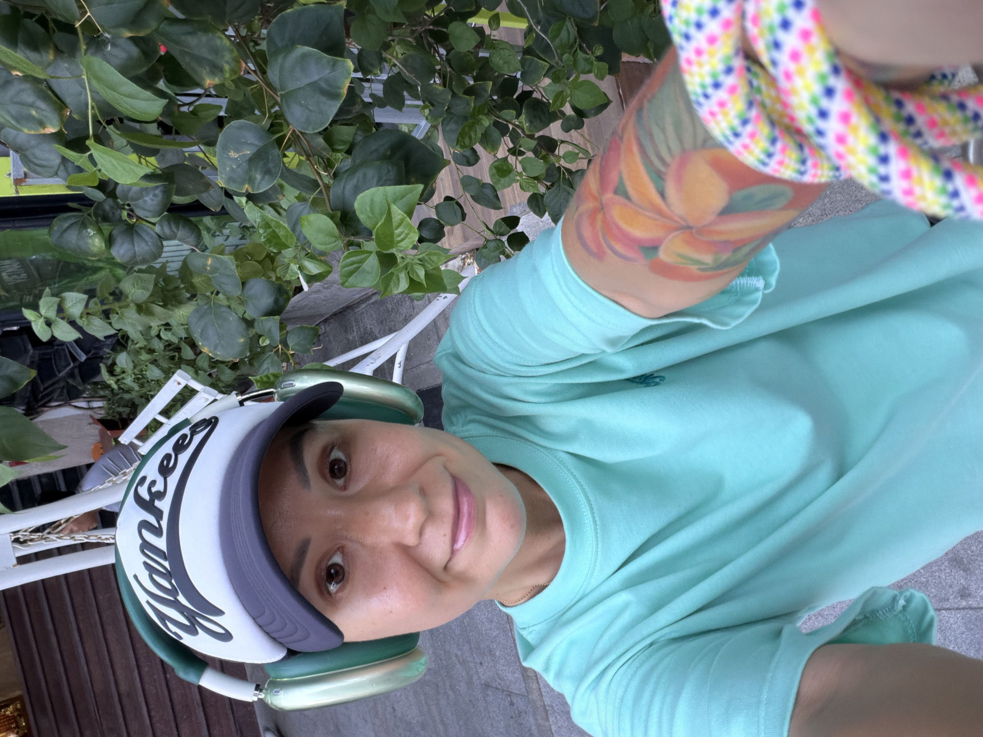 The image shows a woman wearing a teal t-shirt, a Yankees baseball cap, and green headphones. She has a colorful floral tattoo on her left arm and a bright, patterned rope bracelet on her wrist. She is smiling and looking at the camera. The background shows lush green foliage, suggesting an outdoor setting, possibly a garden or a street with plants. A white railing and wooden steps are partially visible, indicating a slightly elevated area or a building structure. The woman appears to be in a public space, and the overall atmosphere is casual and relaxed. The mention of Nha Trang Ward, Vietnam, places the scene in a specific urban environment within Vietnam.
