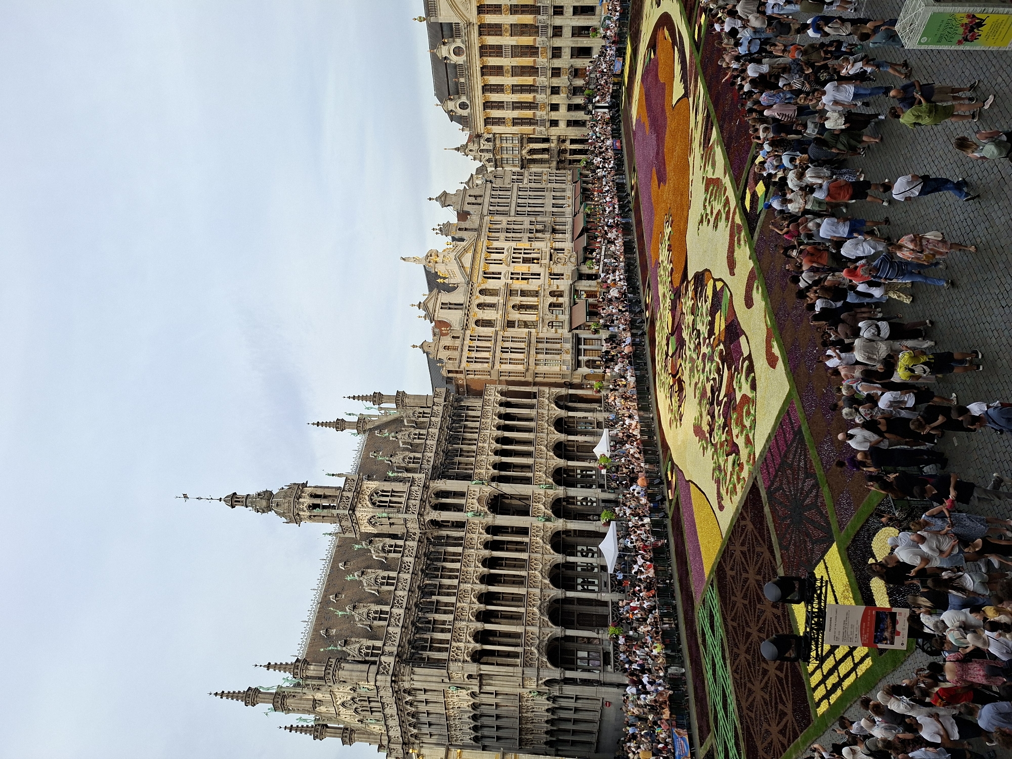 This image captures a vibrant scene at the Grand Place in Brussels, Belgium, during a flower carpet event. The Grand Place, a UNESCO World Heritage site, is renowned for its ornate guildhouses and the magnificent Gothic Town Hall. In this photograph, the square is transformed by an elaborate flower carpet, a biennial event that covers the entire area with intricate floral designs.
The focal point is the sprawling flower carpet, a tapestry of vivid colors and patterns, predominantly featuring hues of purple, orange, yellow, and green, arranged in organic shapes and geometric sections. Thousands of meticulously placed begonias, dahlias, and other flowers form the artwork.
Surrounding the flower carpet, a large crowd of onlookers is visible, cordoning off the display area. People are gathered, observing the floral masterpiece and taking photographs. Barriers and temporary fencing are in place to guide the flow of the visitors and protect the installation.
The background is dominated by the stunning architecture of the Grand Place. To the left, the imposing Gothic facade of the Town Hall, with its detailed spires and arched windows, stands prominently. To the right and across the square, the elegant facades of the guildhouses, with their golden embellishments and varied architectural styles, create a picturesque urban backdrop.
In the foreground, the cobblestone pavement of the Grand Place is visible, with more people milling about. Several floodlights on stands are positioned near the flower carpet, suggesting preparations for an evening event or enhanced viewing. A sign with event information is also visible. The sky above is overcast, providing diffused light that highlights the colors of the flower carpet. The overall atmosphere appears festive and awe-inspiring, drawing a considerable number of visitors to witness this unique spectacle.