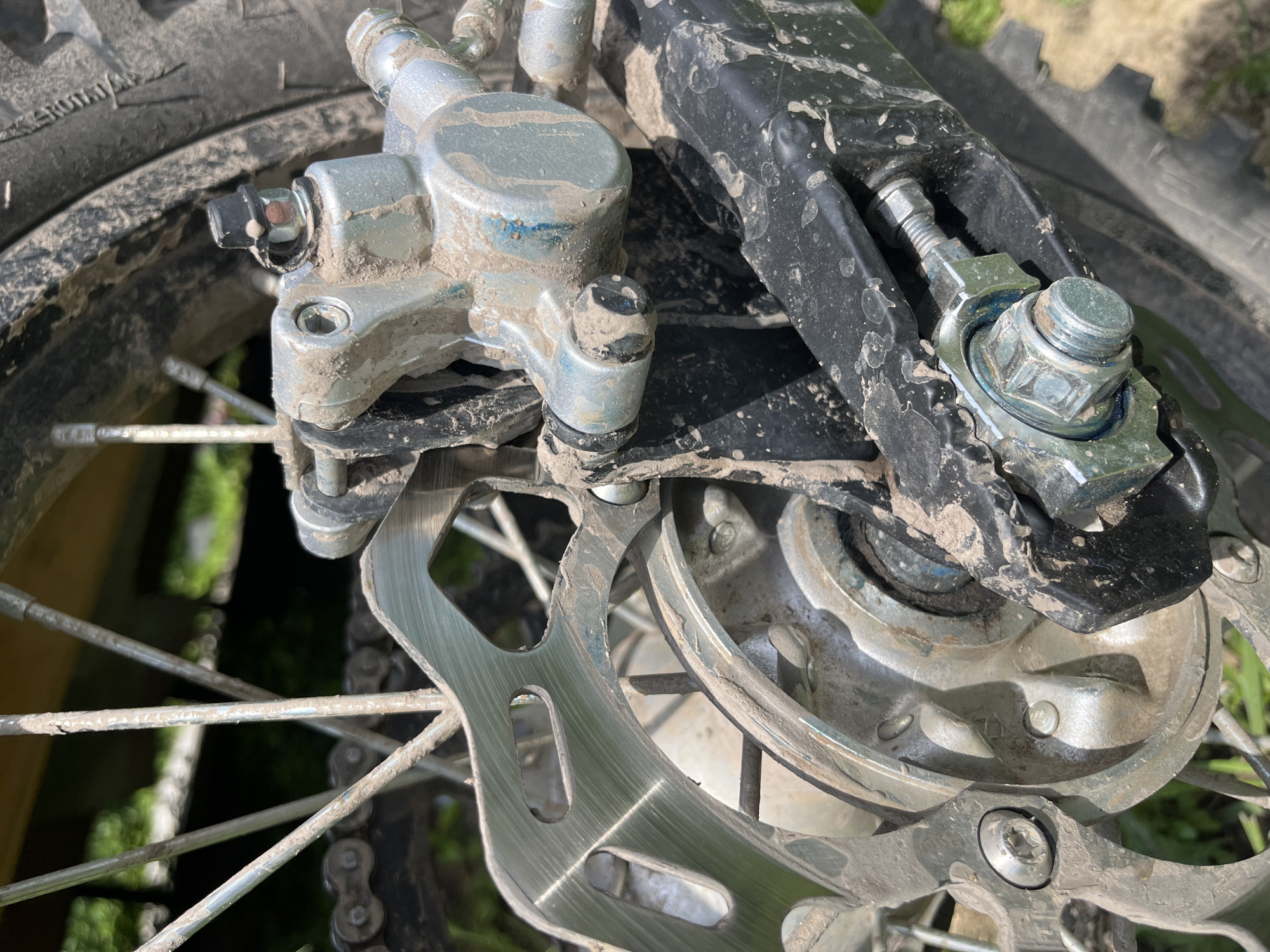 The image displays a close-up, detailed view of the rear wheel brake system of a dirt bike, heavily covered in mud. The scene is outdoors, with blurred green foliage visible in the background, suggesting a natural or unpaved setting. The brake disc, caliper, and chain are all coated in a thick layer of dirt. The tire treads are also visible and caked with mud. A hexagonal bolt and nut securing a part of the swingarm are prominent in the lower right of the frame, also showing signs of mud accumulation. The overall condition of the components suggests recent use in off-road conditions. No people or specific identifiable landmarks are present in the image, and there are no contextual clues to indicate its location within Vasilyevo, Russia.