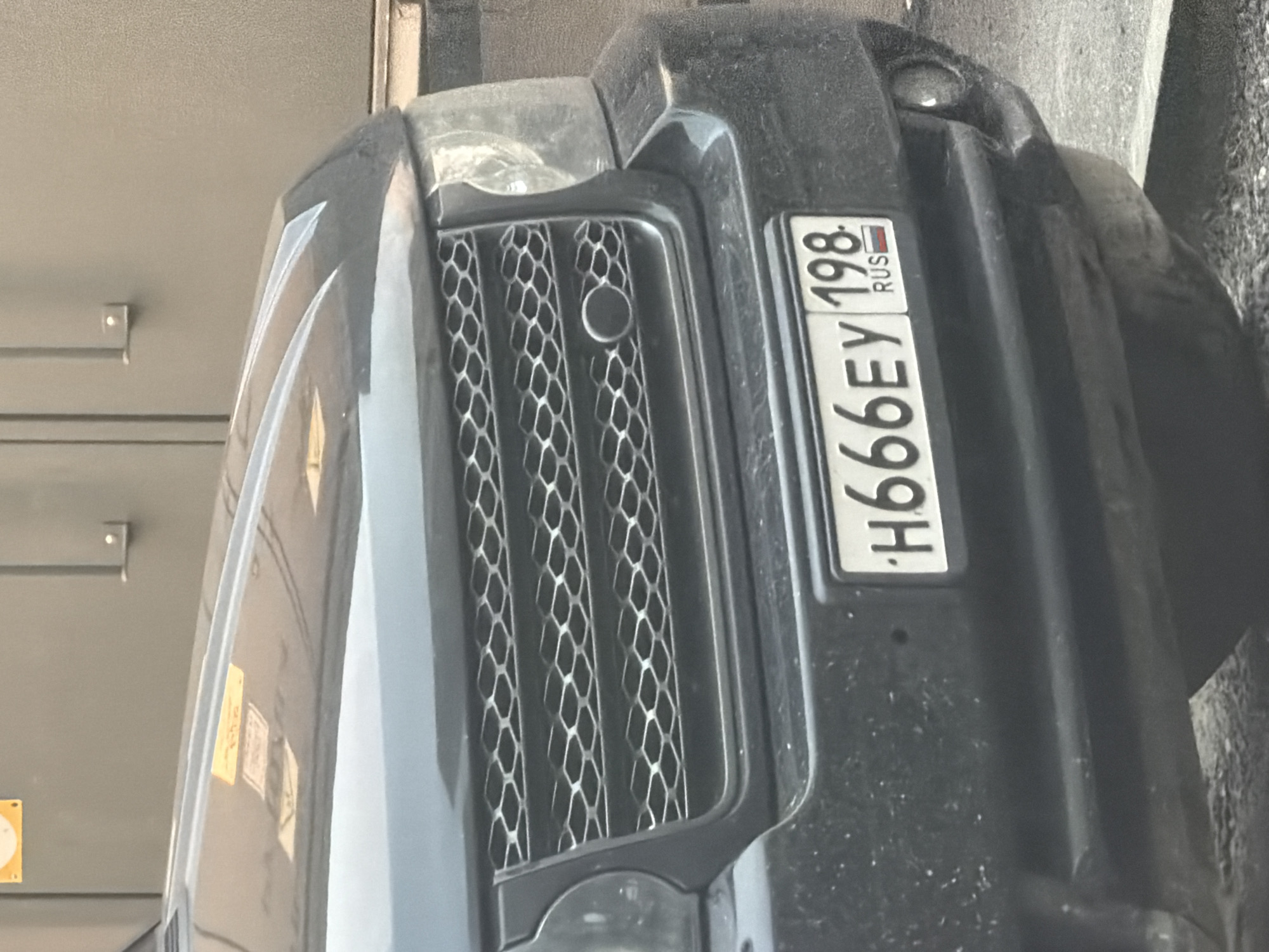 The image shows a close-up of the front bumper and grille of a dark-colored SUV, possibly a Range Rover, parked outdoors. The vehicle is dusty. A license plate with white characters on a black background is clearly visible. The license plate reads "H666EY 198 RUS", with the "RUS" designation and Russian flag on the right side. The surrounding environment appears to be an urban setting, with a grey wall and a hint of pavement. There are no people or other vehicles visible in the frame.