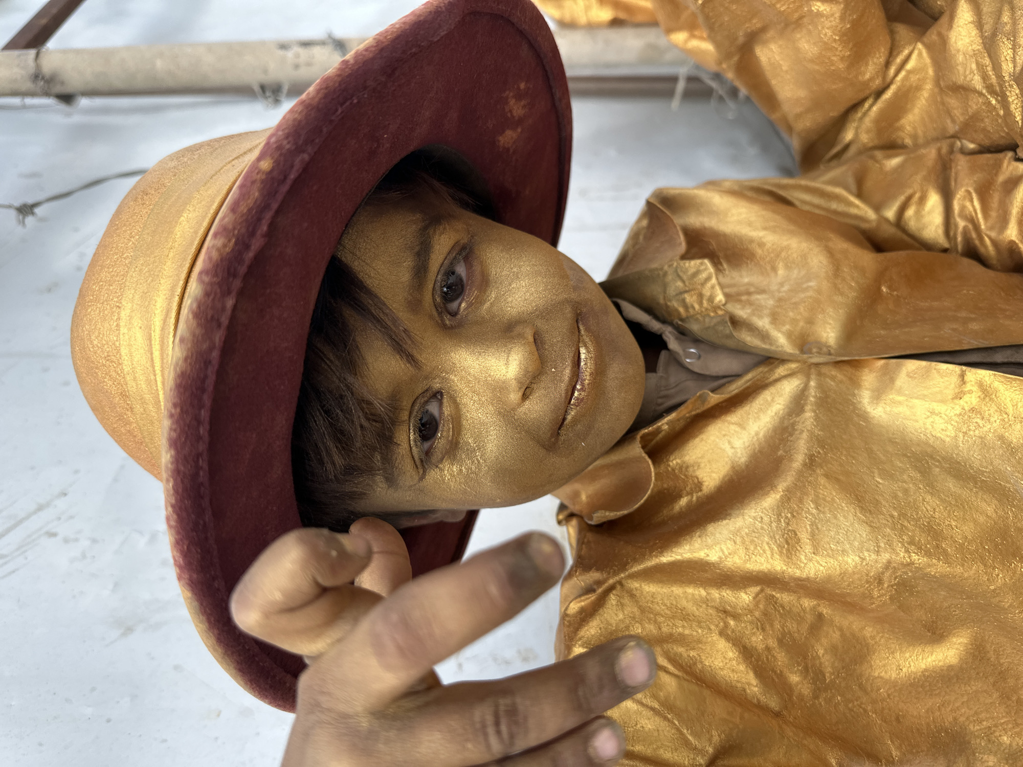 A young person with gold-painted skin is wearing a hat with a gold crown and a maroon brim. The person is wearing a golden jacket. The background is a white wall. It is unknown if the location is in the city of Quetta City Tehsil, Pakistan.