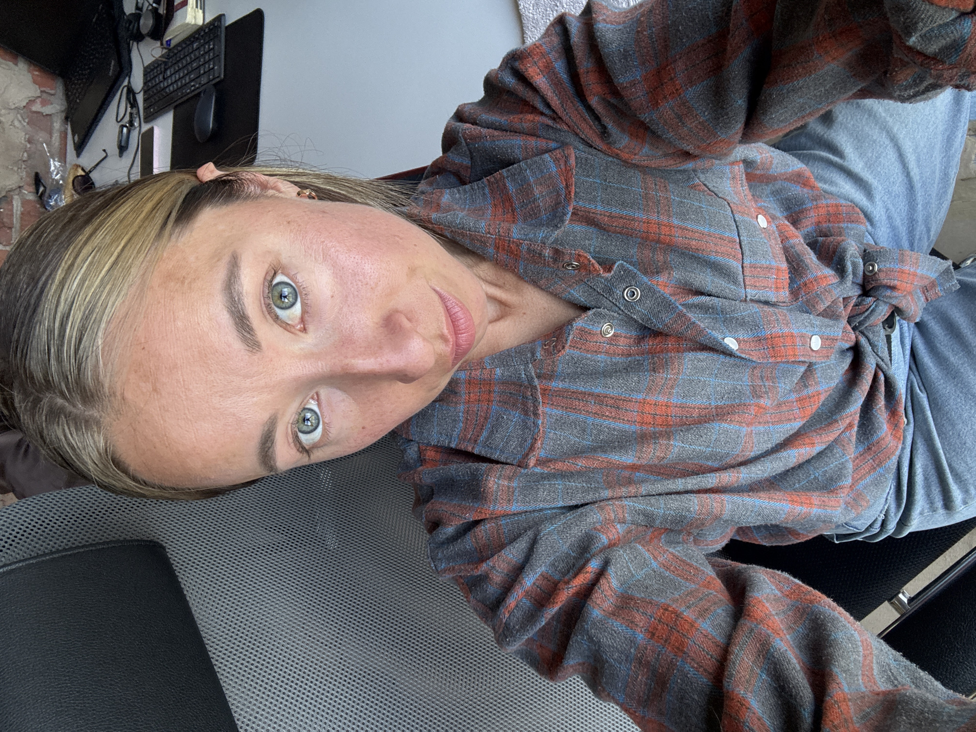 A selfie of a light-skinned woman with blonde hair and green eyes is taken at an extreme upward angle. She is wearing a plaid flannel shirt with orange and blue stripes and light blue jeans. She is seated in a gray mesh chair. A desk with a keyboard, mouse, and laptop is visible in the background. The background also includes a brick wall and a window. The scene is likely indoors, but no information indicates the location is in Chaikovsky, Russia.
