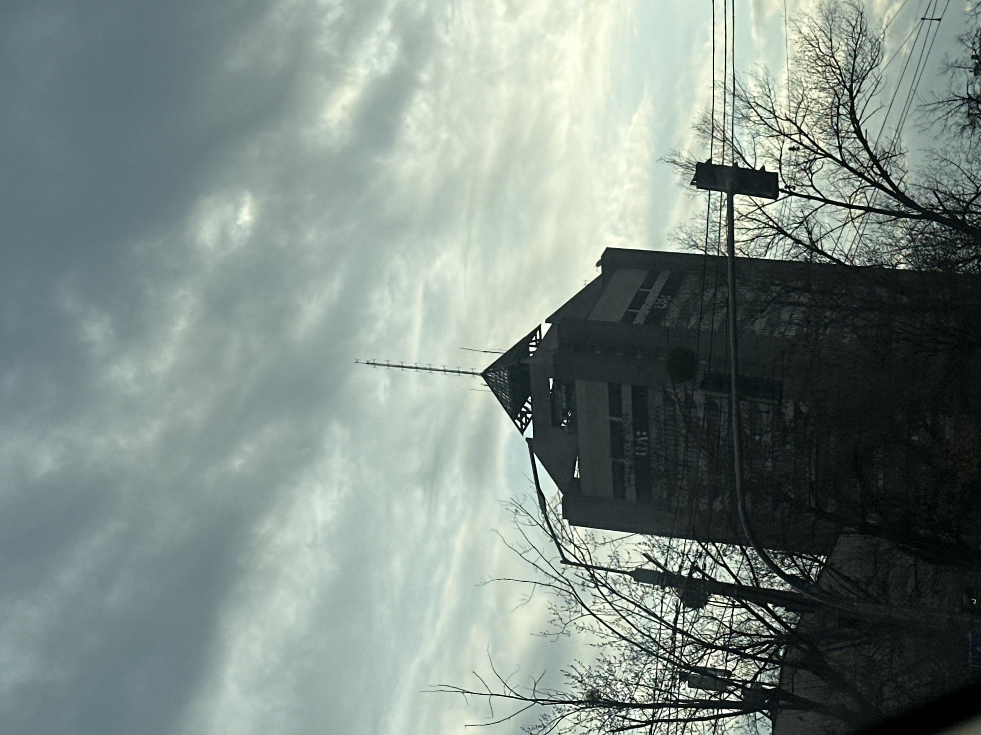 The image captures the silhouette of the abandoned Hotel Parus in Kyiv, Ukraine, against a cloudy sky. The pyramid-shaped building, featuring a prominent antenna, appears deteriorated and unoccupied. Leafless trees and utility poles obstruct parts of the structure, emphasizing the starkness of the urban landscape. The scene is devoid of people or visible activity.