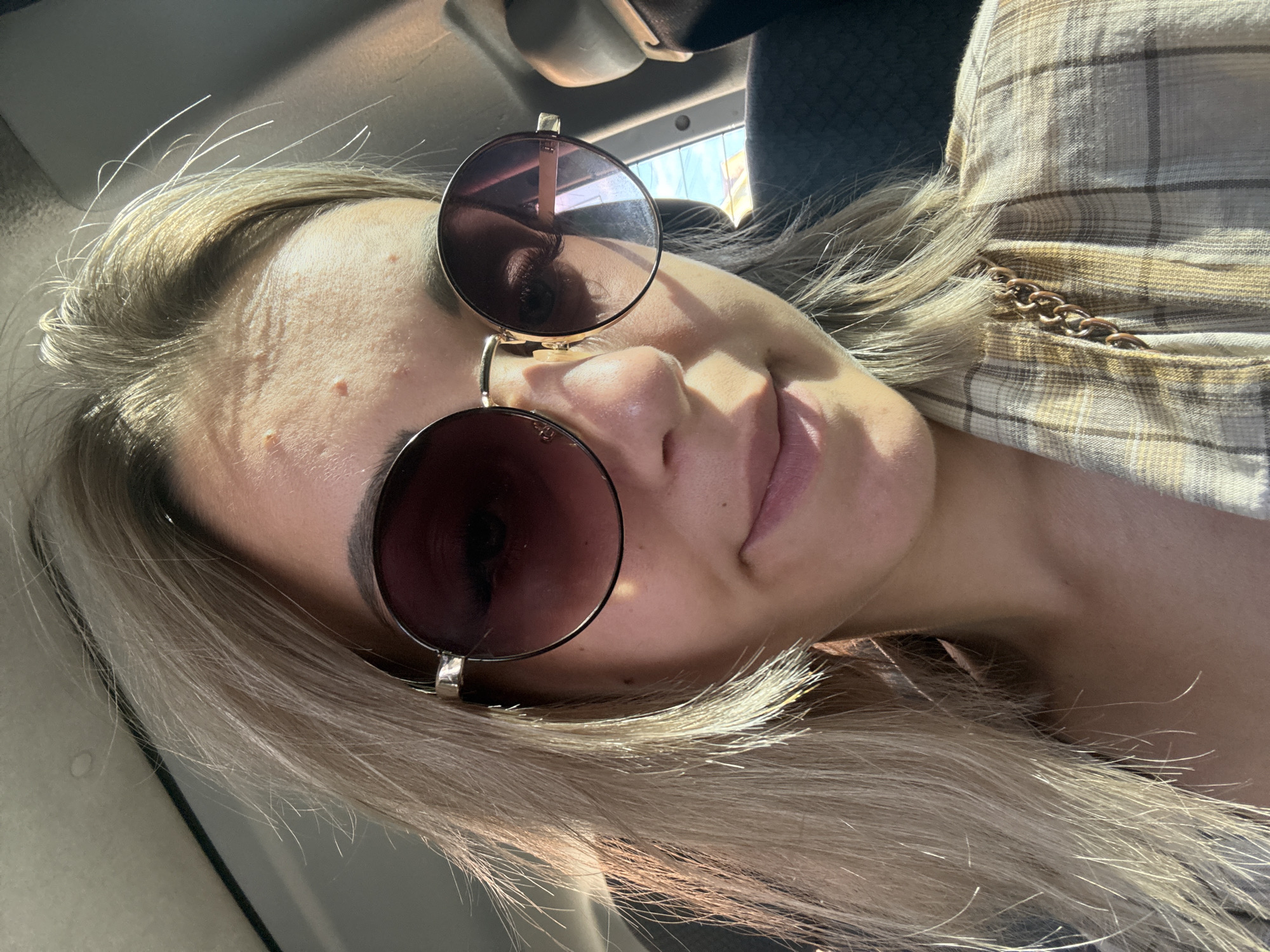 A young woman with blonde hair and sunglasses is in the back seat of a vehicle. She is wearing a plaid jacket with a gold chain. The lighting suggests it is daytime, and the background appears to be a city street, possibly in Yuzhno-Sakhalinsk, Russia, given the context.