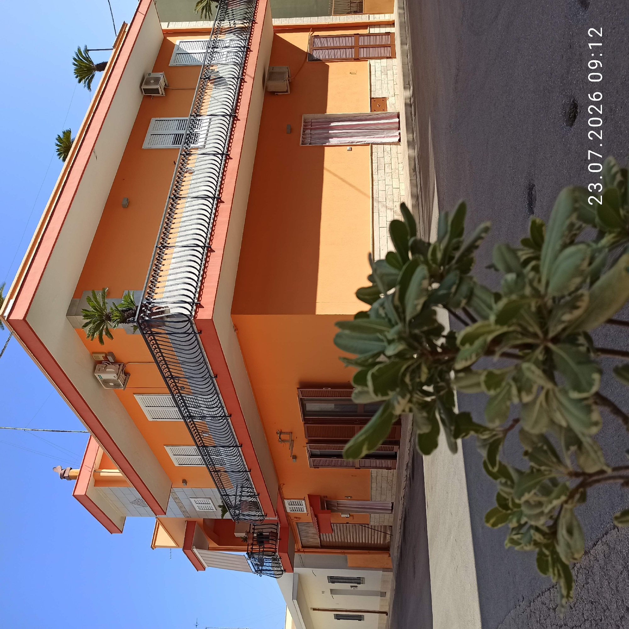 The image shows a corner of an orange multi-story building in Bitonto, Italy, on a clear sunny day. The building has white-framed windows with shutters and a black ornate balcony railing. There are air conditioning units mounted on the exterior walls, and potted plants are visible on the balcony and rooftop. The ground floor features storefronts with closed shutters and a doorway with striped curtains. The street in front of the building is paved, and a out-of-focus plant is in the foreground. The timestamp "23.07.2026 09:12" is visible in the lower right corner.