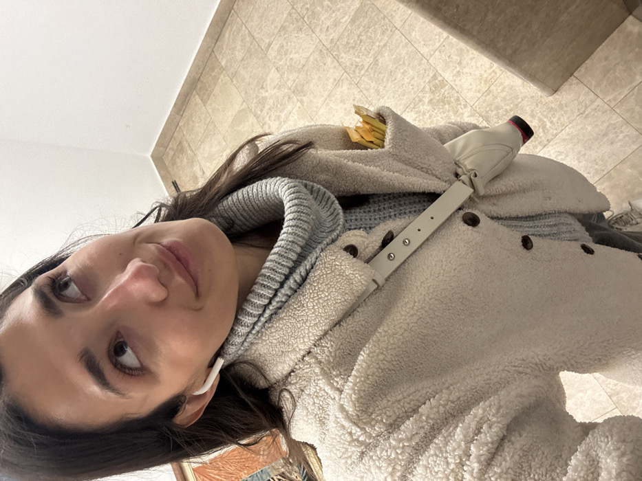 A young woman with brown hair and eyes looks at the camera, wearing an AirPod in her left ear. She has on a gray sweater and a white, fuzzy jacket. A white purse strap with silver grommets goes across her body. The purse is visible over her right shoulder, filled with yellow objects. The background features tile flooring and a wall.