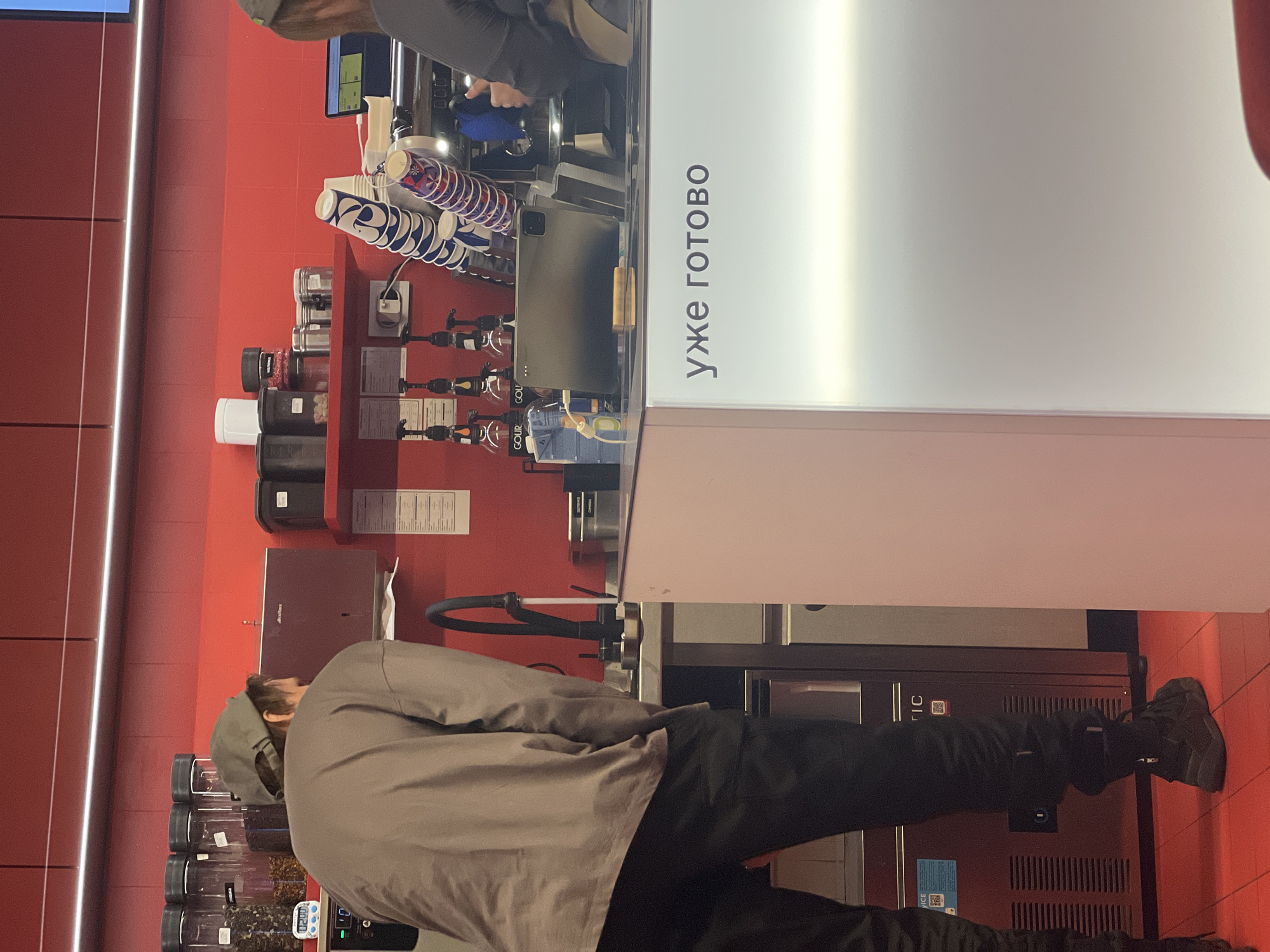 The image, taken indoors and oriented vertically (rotated 90 degrees clockwise), depicts a self-service beverage station or a small cafe counter, likely within a commercial setting.
In the foreground, a person is seen from behind, wearing a gray long-sleeved shirt and a dark baseball cap. They are facing a counter, seemingly interacting with or waiting at the station. Their lower body is clad in dark pants and dark sneakers.
The main focus is the counter area, which features various beverage-related items. On the left side of the counter, numerous disposable cups, some with blue and red patterns, are stacked or held in dispensers. Several dispensers with spigots are arranged, possibly for syrups or flavorings. Jars and containers filled with ingredients, perhaps toppings or coffee beans, are also visible. A tablet device, resembling an iPad Pro, is placed on the counter. Behind this counter, another person's arms are partially visible, operating a machine, suggesting they are preparing drinks or stocking the station.
Dominating the right side of the counter is a tall, white vertical panel with bold, dark text written in Cyrillic. The text reads "УЖЕ ГОТОВО", which translates from Russian to "ALREADY READY" or "IT'S READY NOW". This sign likely indicates that an order is complete or a drink is prepared for collection.
The background consists of a bright red tiled wall, suggesting a modern and clean aesthetic. Overhead, there's bright artificial lighting, causing reflections on the white panel. The overall scene indicates a bustling, efficient setup for dispensing beverages. Given the location context of Khimki, Russia, the Russian text is consistent with the geographical setting.