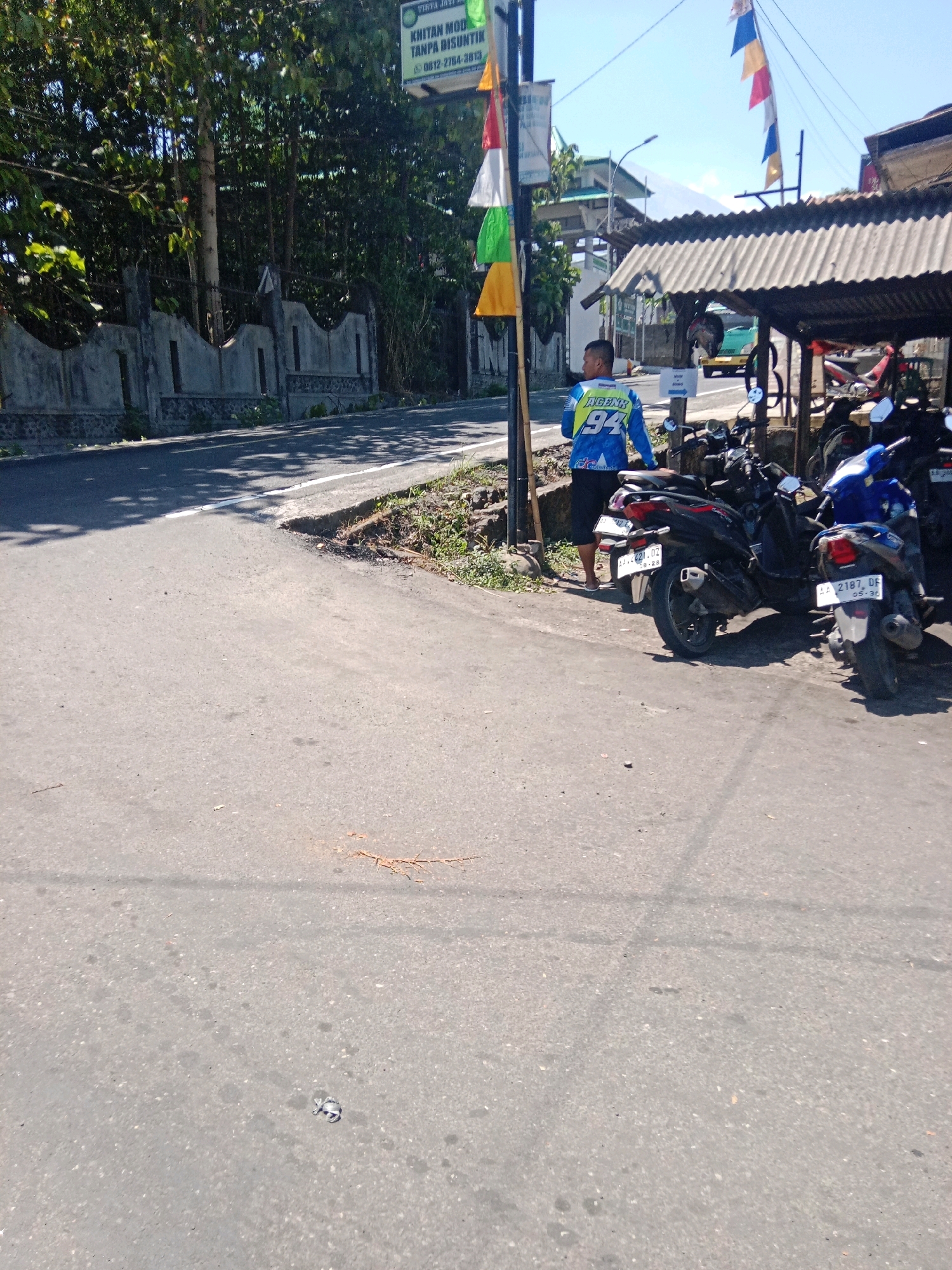 In the city of Kertek, Indonesia, a man wearing a blue and yellow jacket with the number "94" visible stands on the side of an asphalt road, facing away from the camera. He is wearing dark pants and white flip-flops. Several motorcycles are parked in front of a building with a corrugated metal roof. The building appears to be a shop or a small business. In the background, trees and a concrete wall are visible. Colorful flags are strung on a pole near the building. The road is somewhat cracked and appears to be well-trafficked. The sky is clear and blue, suggesting it is daytime.