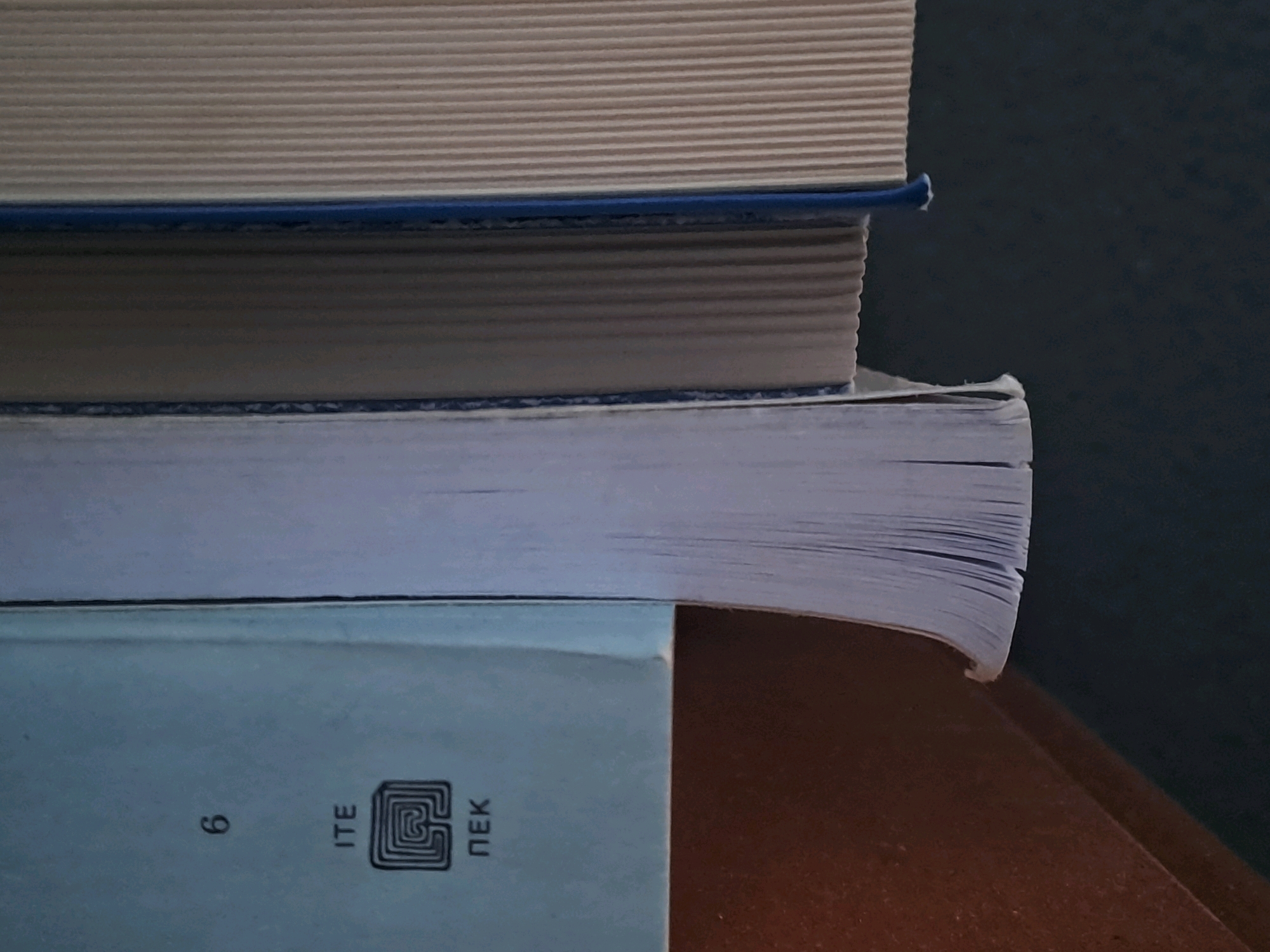 A close-up, horizontally oriented photograph shows a stack of four books. The books are stacked vertically. The top two books have light tan covers with a ribbed texture, and blue binding at the spine. The third book from the top has a white cover and a slightly fanned out edge. The bottom book has a light blue cover with white text. The text includes the Cyrillic characters "6", "ITE", and "ПЕК". A black, square logo with a maze-like pattern is also visible on the bottom book. The books are resting on a wooden surface that is partially visible on the right side of the frame. The background is a dark, textured surface. The image is well-lit, with the light source appearing to be from the left, casting subtle shadows. There are no people or animals present in the image. The scene is an interior setting, likely a home or office. The location of Kolchiko, Greece is not relevant to this image.
