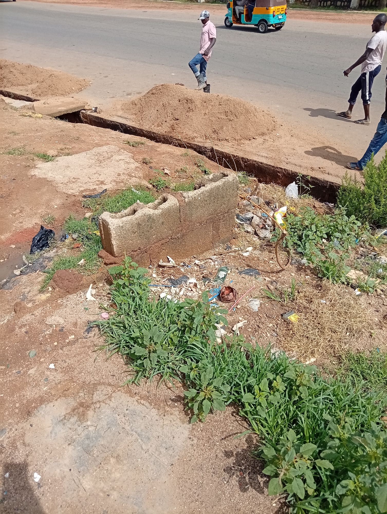 The image depicts a roadside scene in Jos, Nigeria. A trench has been dug alongside the road, bordered by piles of sand. Debris and weeds are present around the trench. Two stacked cinder blocks are positioned near the trench. Three people are visible: one is walking near a yellow and blue vehicle on the road, while two others are walking closer to the scene. The setting appears to be urban, with a paved road and signs of construction or infrastructure work.