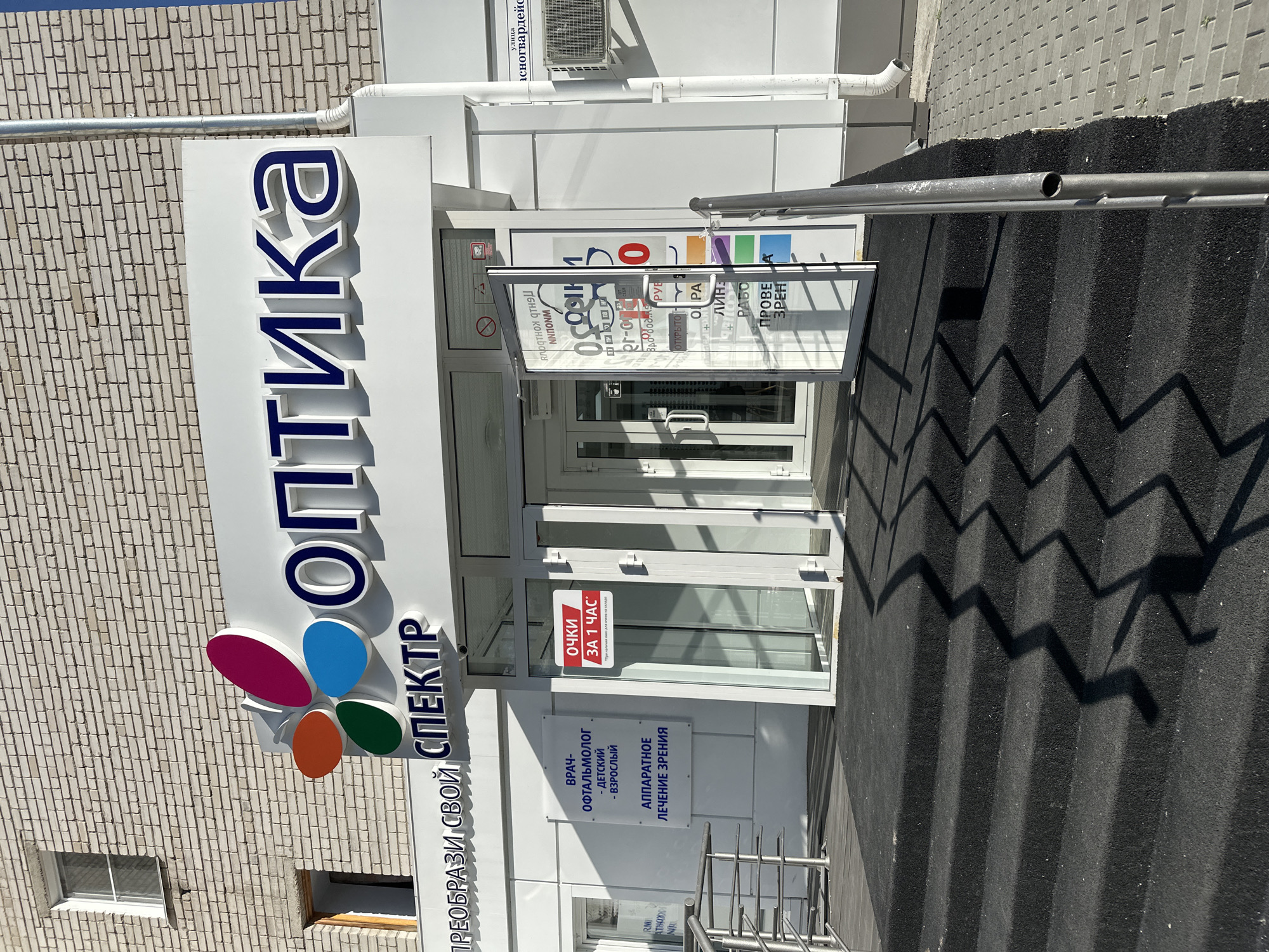 This media file captures the exterior of an optics store in Novoaltaysk, Russia. The prominent signage features the word "ОПТИКА" (Optika) in large, blue, three-dimensional letters on a white background, with a colorful logo resembling a flower or butterfly below it. To the right of the main entrance, a glass door is propped open, revealing the interior and more signage.
Visible text on the exterior includes "ПРЕОБРАЗИ СВОЙ СПЕКТР" (Transform Your Spectrum), indicating the store's name or slogan. A red and white sticker on the glass door advertises "Очки ЗА 1 ЧАС" (Glasses in 1 Hour), suggesting a quick service. Inside the store, further text is visible on posters, including "ВРАЧ-ОФТАЛЬМОЛОГ" (Ophthalmologist), "ДЕТСКИЙ - ВЗРОСЛЫЙ" (Child - Adult), and "АППАРАТНОЕ ЛЕЧЕНИЕ ЗРЕНИЯ" (Apparatus Vision Treatment), detailing the services offered.
The building's facade is made of light-colored brick on the left and white siding on the right. The entrance is modern with glass doors and a smaller glass window to the left. In front of the store, there is a textured, dark-colored paved area with a metal railing. The scene is illuminated by bright sunlight, casting sharp shadows of the railing onto the pavement, suggesting a clear, sunny day. The overall impression is a well-maintained, professional establishment ready to serve its customers. No people are visible in the frame.