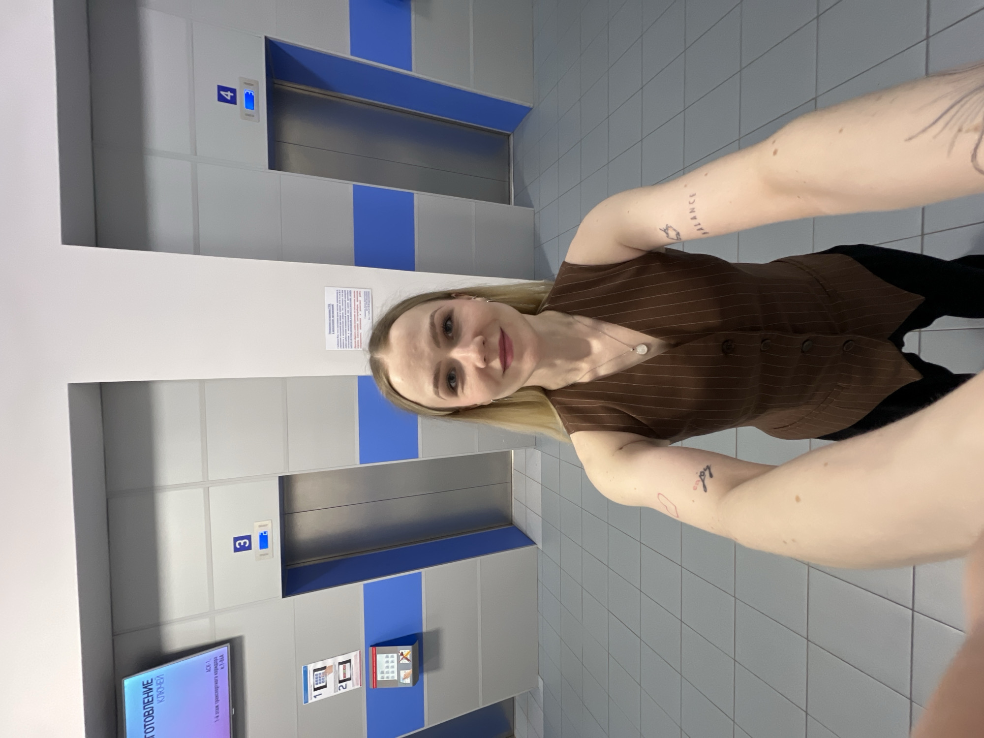 The image captures a selfie of a woman in a modern elevator lobby. The lobby has two elevator doors with blue accents. The elevator buttons indicate access to floors "3" and "4". A digital screen displays text in Russian indicating information for a "transport corridor". There are also informational posters near the elevator doors. The woman is wearing a brown, pinstriped vest, and has visible tattoos on her arms, with some text that appears to read "balance". The presence of Russian text and general appearance suggest the location may be in Khimki, Russia.
