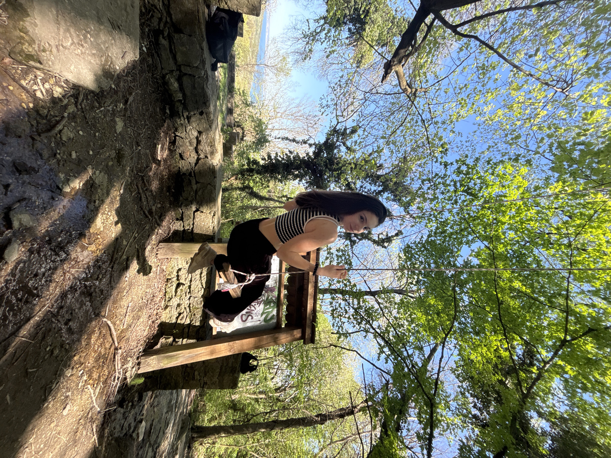 The image depicts a woman sitting on a small wooden structure, possibly a dilapidated gazebo or shelter, within a wooded area. The woman has dark hair and is wearing a striped top and black pants. She is holding a thin stick or string. The trees around her are lush and green, suggesting a park or forested area. In the background, a body of water (possibly the Black Sea) and the sky are visible. A stone wall or structure is present on the left side of the frame, and what appears to be a bag sits atop this structure. Based on the location details, the scene is likely situated in or near one of the parks or natural areas in Varna, Bulgaria, possibly Primorski Park.