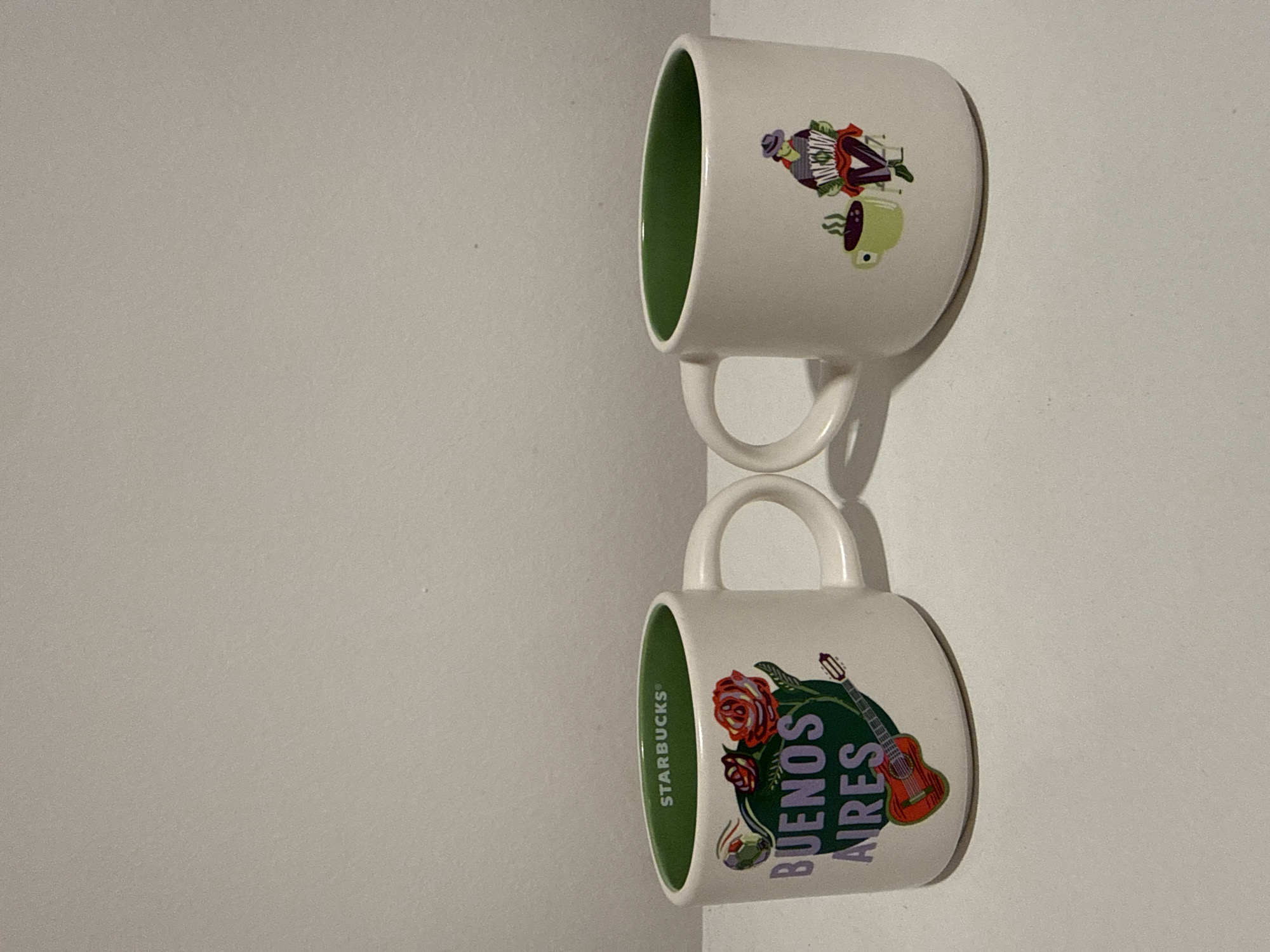 The image displays two white ceramic Starbucks mugs with vibrant green interiors, hanging against a plain, light-colored wall. The photo is rotated 90 degrees clockwise, so the mugs appear to be hanging vertically with their handles facing left.
The top mug features a colorful cartoon illustration. On its side, there is a figure dressed in a hat and striped clothing, playing an accordion. Beside this character, there is a stylized green mug or cup with steam rising from it. The overall design evokes themes possibly related to music or cultural elements like tango.
The bottom mug showcases a design that explicitly identifies its location. The words "BUENOS AIRES" are prominently displayed in purple text on a dark green, somewhat organic-shaped background. Accompanying the text are illustrations of red roses and a classical acoustic guitar. Vertically printed along the inner rim of this mug, the "STARBUCKS®" logo is clearly visible in white text against the green interior.
Both mugs have white handles. The background is simple, suggesting they are being stored or displayed. The designs on both mugs, particularly the "BUENOS AIRES" text and the accordion player, strongly indicate a connection to Buenos Aires, Argentina, aligning with the provided location context. No activity is taking place; the mugs are stationary objects.