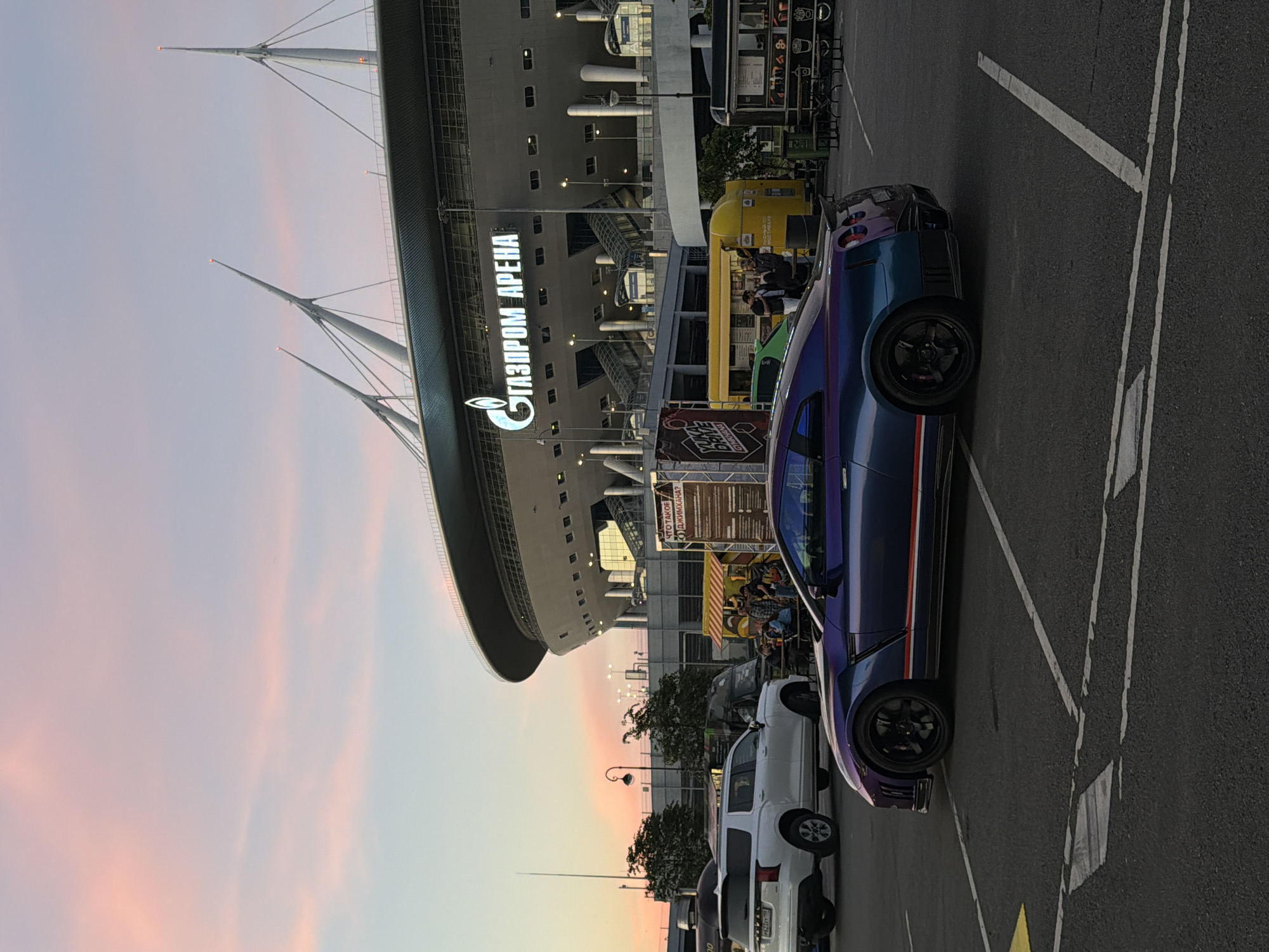 The image captures a street scene with a modern stadium in the background, identified by the prominent "ГАЗПРОМ ARENA" signage. The sky displays a soft pink and blue gradient, indicative of either dawn or dusk. In the foreground, a dark blue sports car with iridescent purple accents is parked. To the left, a white SUV is also parked. Behind the vehicles, several food trucks are visible, with people gathered around them, suggesting a public event or gathering. The overall environment appears to be an outdoor paved area, likely a parking lot or public square, with streetlights and trees present.