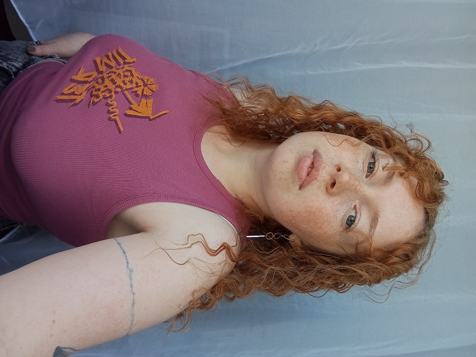 A red-haired woman with freckles is lying on a light blue surface, possibly a bed or fabric. She is wearing a mauve-colored tank top with an orange graphic design. She has an earring on her left ear. A tattoo is visible on her left arm. The location is not identifiable as Yurgamysh, Russia, based on the image provided.