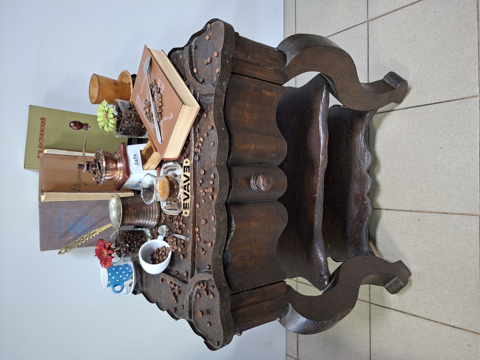 A still life scene is set on an antique wooden table. The table has intricate carvings and a lower shelf. Scattered across the table's surface are coffee beans.
On the table, several objects are arranged:
- A blue and white polka dot teacup and saucer.
- A white teacup, tipped over, with coffee beans spilling out, alongside a spoon.
- A glass containing coffee beans and a red flower.
- A copper coffee pot (cezve).
- A small glass filled with dark liquid, presumably espresso.
- A metal coffee grinder.
- A wooden box with ground coffee.
- A brown book with coffee beans scattered on its cover, and a spoon resting on top.
- A glass cup filled with coffee beans and a green flower.
- A wooden cup or holder with a saucer.
- Two books are visible in the background, leaning against each other.
The scene appears to be indoors, with a light-colored wall behind the table and tiled flooring in the foreground. The overall aesthetic suggests a focus on coffee and perhaps a vintage or artistic theme. The objects are arranged in a deliberate composition, creating a visually appealing display. There are no people or animals present in the image, and no specific location in Chișinău, Moldova can be identified from the provided visual information.