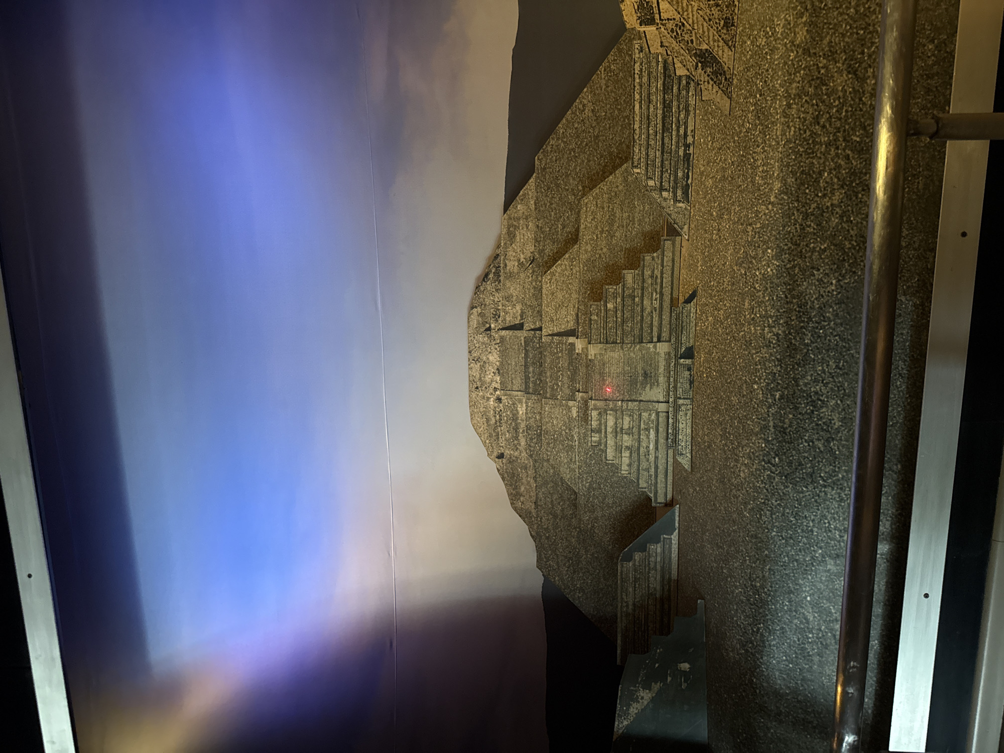 The image displays a detailed model or exhibit of a large, stepped pyramid or ancient structure, likely within a museum or exhibition setting in Mexico City, Mexico. The structure, which resembles a Mesoamerican pyramid with multiple terraces and a rough, granular texture suggesting stone construction, dominates the right side of the frame. It is illuminated by warm, artificial light, casting defined shadows that accentuate its architectural steps and form. A small red light dot is visible near the top center of the pyramid.
To the left, a vibrant, gradient backdrop simulates a sky, transitioning from deep blue and purple hues at the bottom to a lighter, hazy greyish-blue at the top, suggesting either dusk, night, or an abstract lighting effect. A faint, thin white line stretches horizontally across this simulated sky, possibly a contrail or a mark on the display surface.
On the far right, a dark metal handrail or support bar runs vertically, extending beyond the top and bottom edges of the frame, indicating the viewer's proximity to an exhibit. Another horizontal metal bar is attached to it further right. The overall impression is an indoor, immersive display designed to showcase the ancient architecture characteristic of the region. There is no visible text or identifiable human activity.