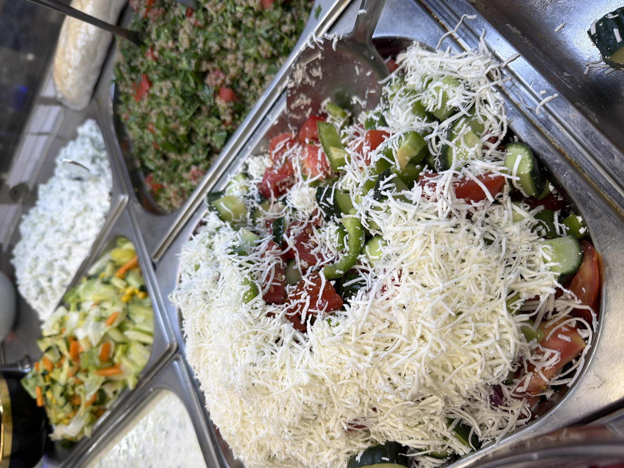 The image depicts a close-up view of several food service trays filled with various salads, suggesting a buffet or deli setting. The primary focus is a large tray overflowing with a chopped salad composed of tomatoes, cucumbers, and green peppers, generously topped with a thick layer of shredded white cheese. In the background, other trays hold different salad varieties: one with a white creamy dressing, another with a green salad containing carrots and corn, and a third filled with a finely chopped grain-based salad, possibly tabbouleh, mixed with parsley and tomatoes. A loaf of bread is visible in the upper right corner. The overall presentation is indicative of a self-serve food establishment. There are no people or animals visible in the image. The lighting appears to be artificial, common in indoor food service environments. The food is presented in stainless steel trays, typical for holding prepared foods.