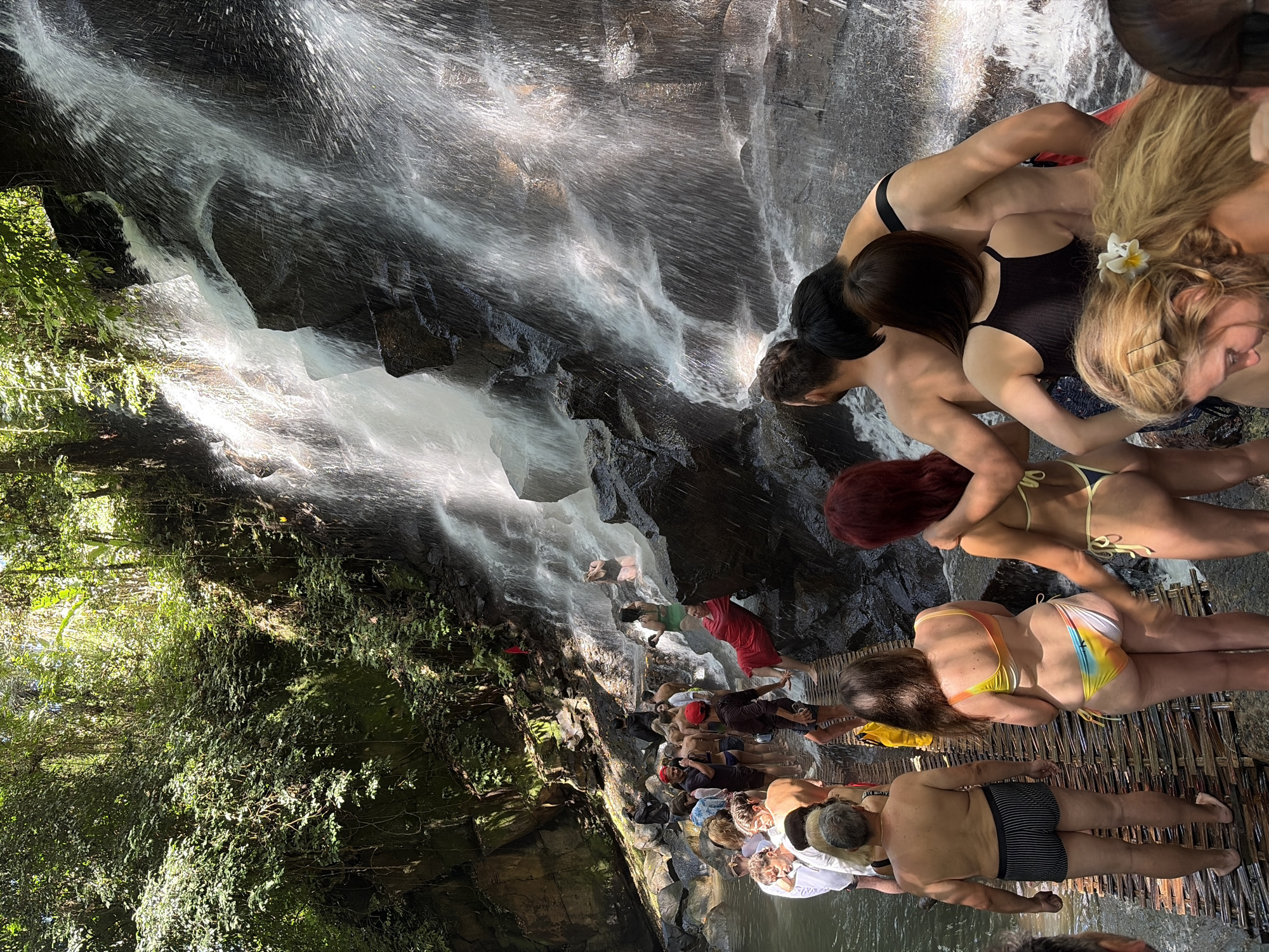 A diverse group of people, both male and female, are gathered around a waterfall in Gianyar, Indonesia. The waterfall cascades down a rocky cliff face, with lush green vegetation surrounding the area. Many individuals are dressed in swimwear, suggesting they are participating in recreational activities at the waterfall. Some are standing on a wooden walkway that leads towards the waterfall, while others are in the water or on the rocks. The atmosphere appears to be lively and social, with people interacting and enjoying the natural scenery.