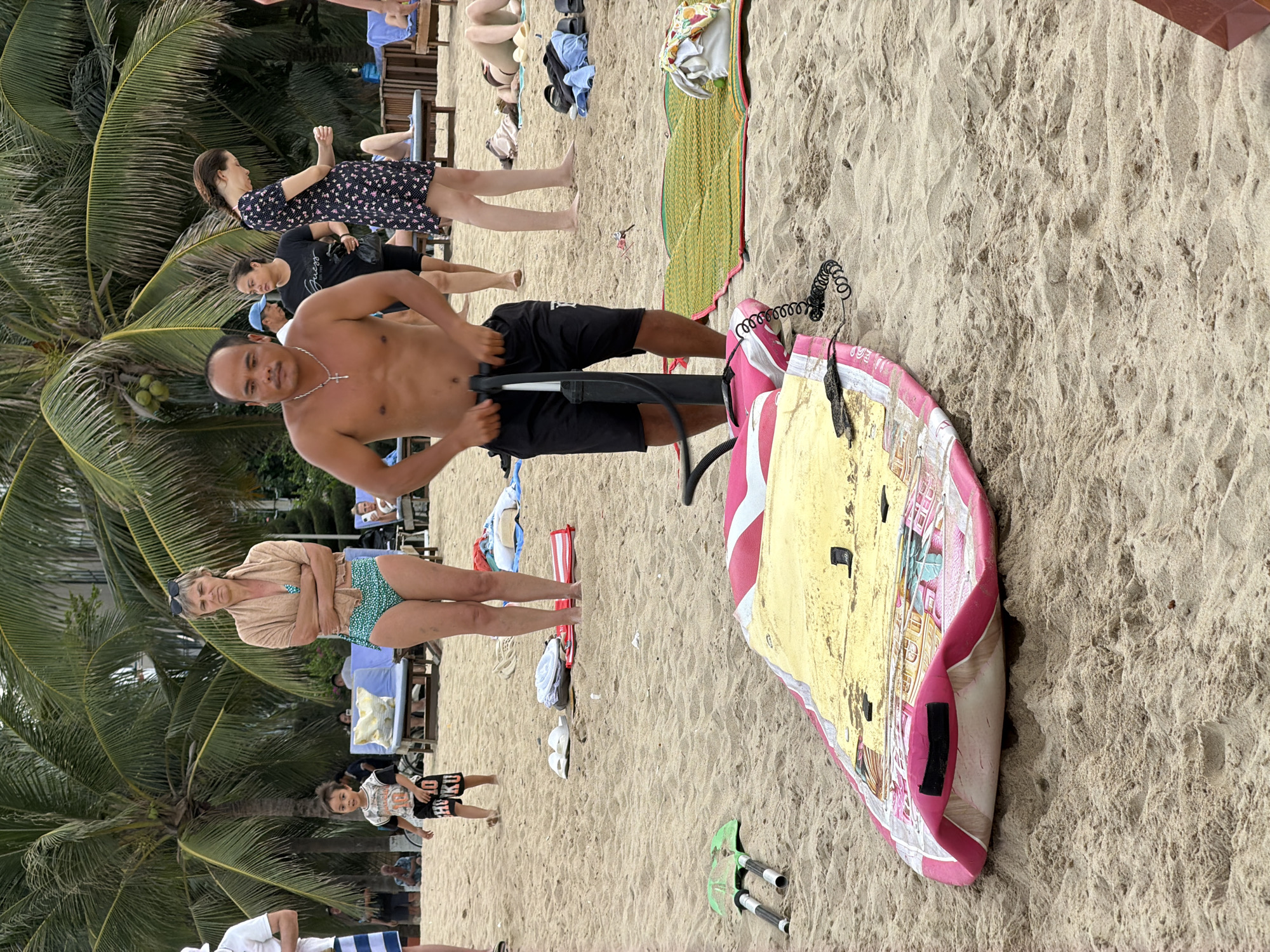 The image depicts a beach scene in Nha Trang Ward, Vietnam, likely a public area with palm trees, sand, and lounge chairs. A man in black shorts is actively using a hand pump to inflate a pink and yellow inflatable board, which lies on the sand in the foreground. Other people are visible in the background, including a child in a t-shirt and shorts, a woman in a patterned dress, and a woman in a bikini and shawl. The environment appears to be a sunny day, with the sand and water being the primary textures. Several objects are scattered on the sand, including a pair of green flippers and a woven mat.