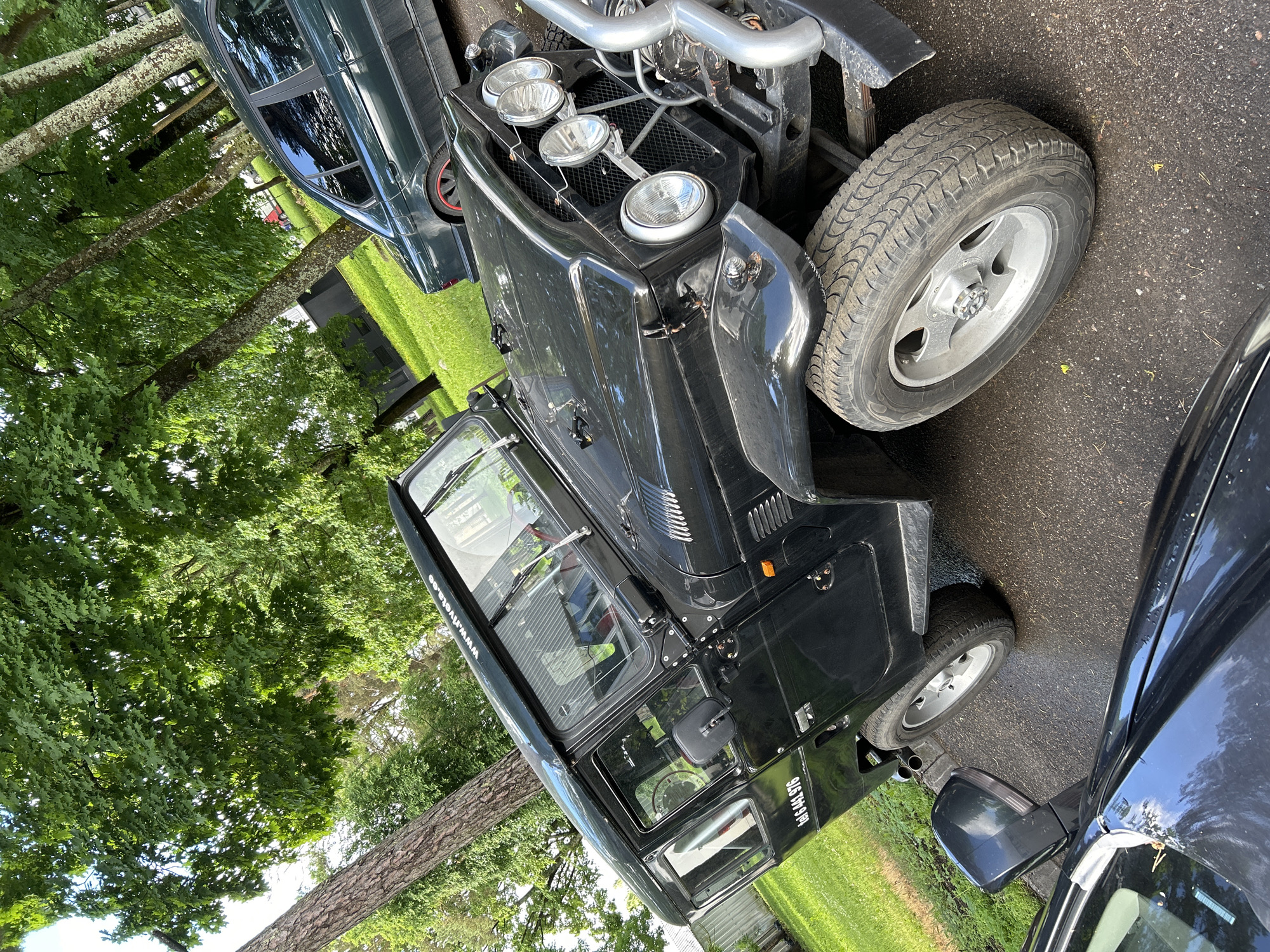 The image shows a black, classic off-road vehicle, likely a Toyota Land Cruiser, parked on an asphalt surface next to a grassy area with trees. The vehicle has a chrome bumper and auxiliary lights on the front. A green sedan is parked behind it. A partial view of a dark-colored car is in the foreground. The background features lush green trees and some buildings. There are no people visible in the image. The license plate of the off-road vehicle reads "tel 6 441 976". A website address "www.riveta.ee" is visible on the top of the off-road vehicle's windshield.
