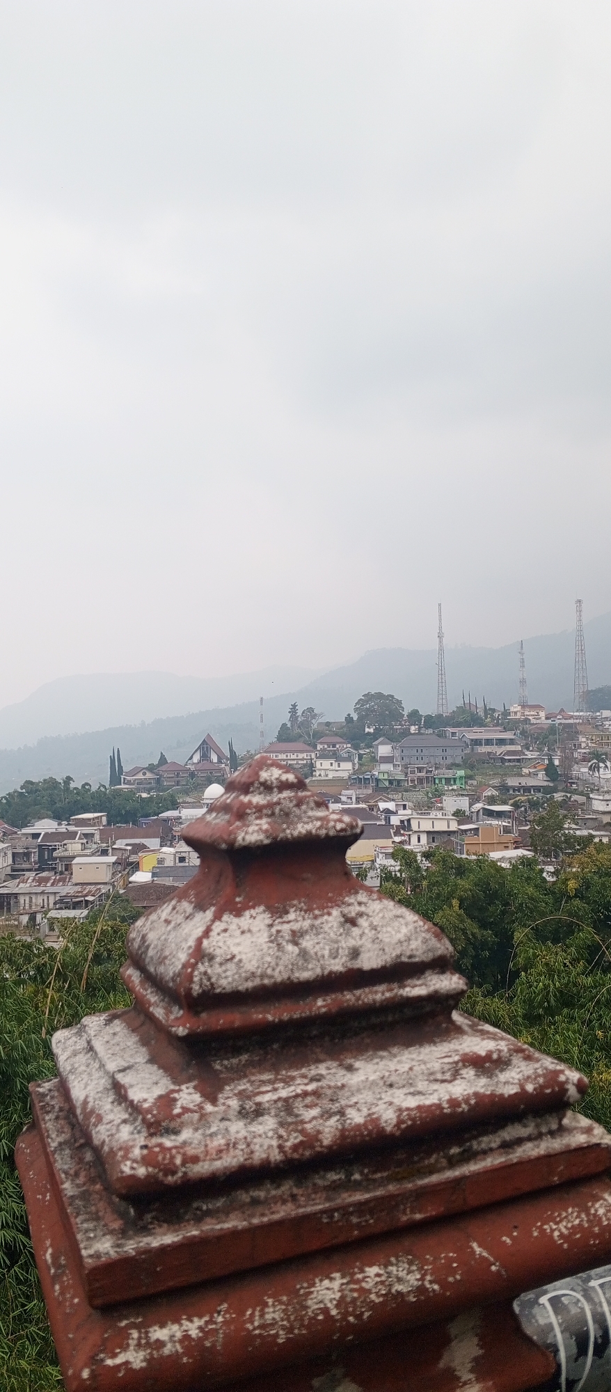 In Sarangan, Indonesia, an overcast sky looms above a densely packed hillside town. The foreground is dominated by a weathered, tiered architectural element, likely part of a temple or monument, made of reddish-brown material with white patches of discoloration. Behind this structure, the town unfolds with a variety of buildings of different colors and sizes, interspersed with green trees and vegetation. Several tall telecommunication towers are visible on the distant hills, piercing the hazy atmosphere. The overall impression is of a sprawling settlement nestled in a mountainous or hilly terrain.