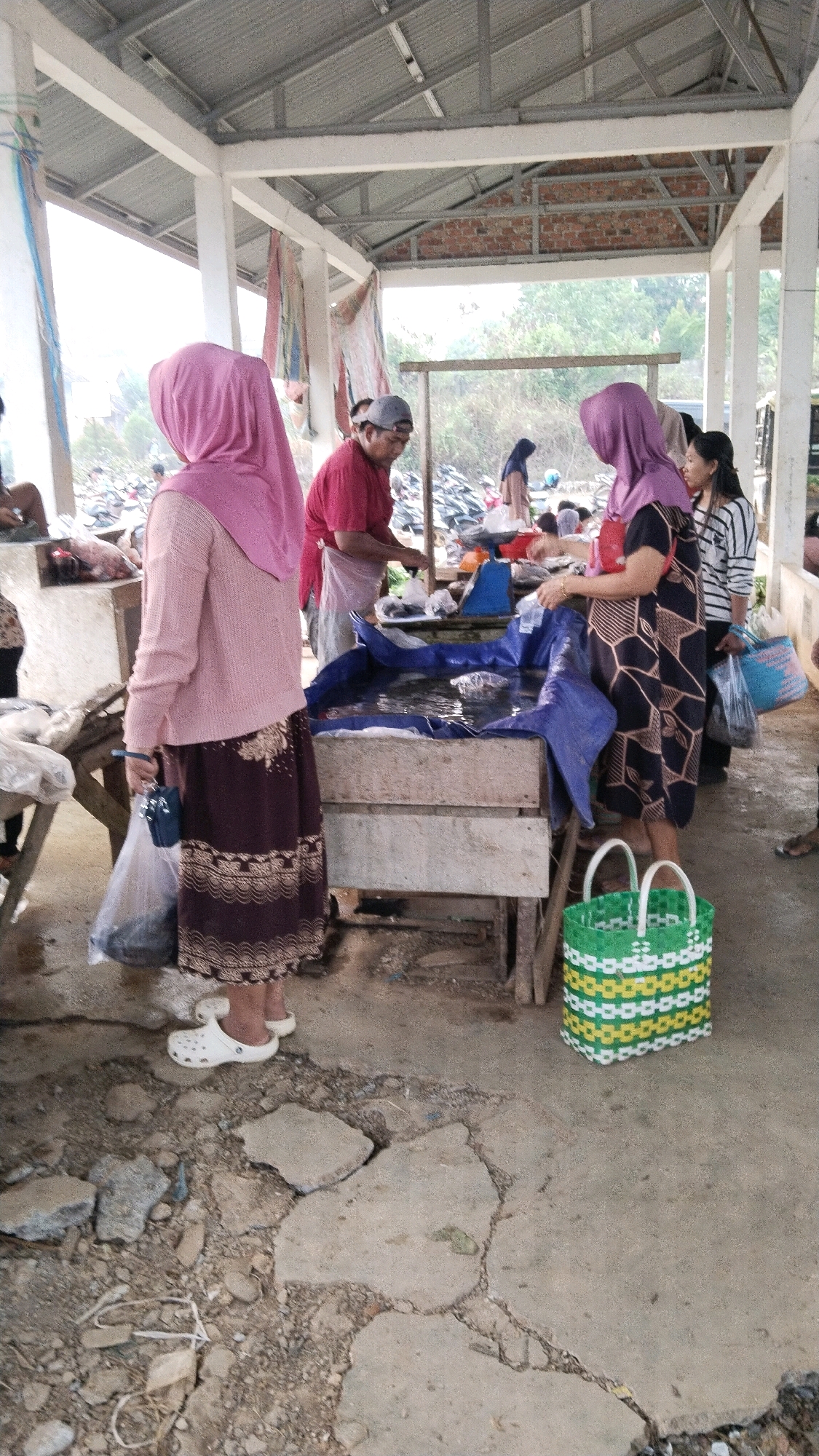 The image depicts an outdoor market scene in Marga Sakti, Indonesia. In the foreground, a woman wearing a pink headscarf and a patterned skirt stands next to a plastic bag filled with goods. She is wearing white clogs. To her right, a green and yellow woven basket sits on the ground. Behind her, a wooden table holds a blue tarp filled with water, likely for displaying fish. A man in a red shirt and grey cap stands behind the table, appearing to be working with the fish. Another woman in a purple headscarf and a black and white patterned dress is standing at the table, holding a red purse. In the background, more people, motorcycles, and a covered structure with a metal roof are visible. The ground is a mixture of concrete and dirt with scattered rocks and debris. The overall impression is of a bustling local marketplace.