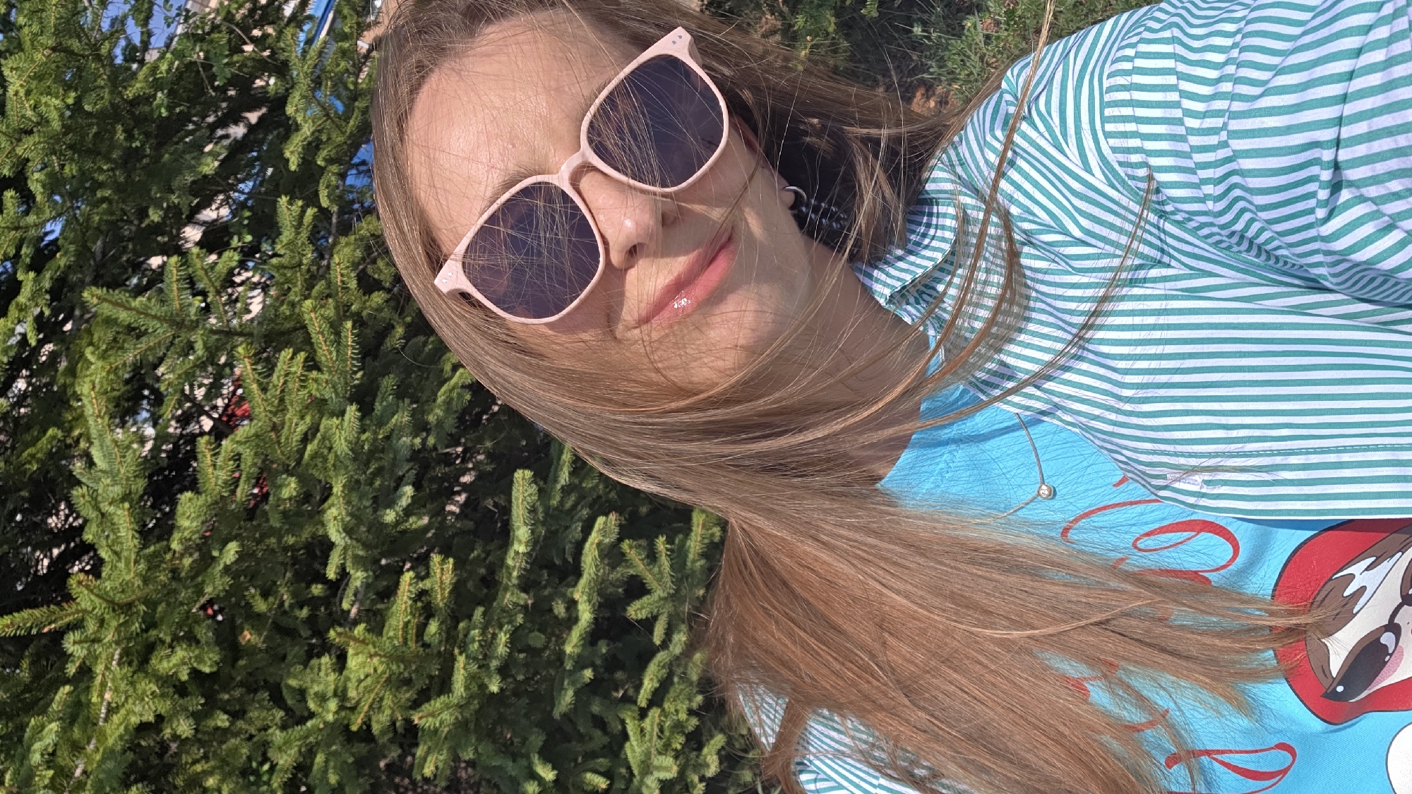 The image shows a young woman with long, light brown hair wearing pink sunglasses and a light blue and white striped shirt. She is standing in front of a lush, green coniferous tree. Part of a light blue graphic t-shirt is visible beneath the striped shirt, featuring red lettering and a cartoon graphic. The location is suspected to be Birobidzhan, Russia, given the request, but there are no specific visual cues in the image to confirm it. The photograph appears to have been taken outdoors in daylight.