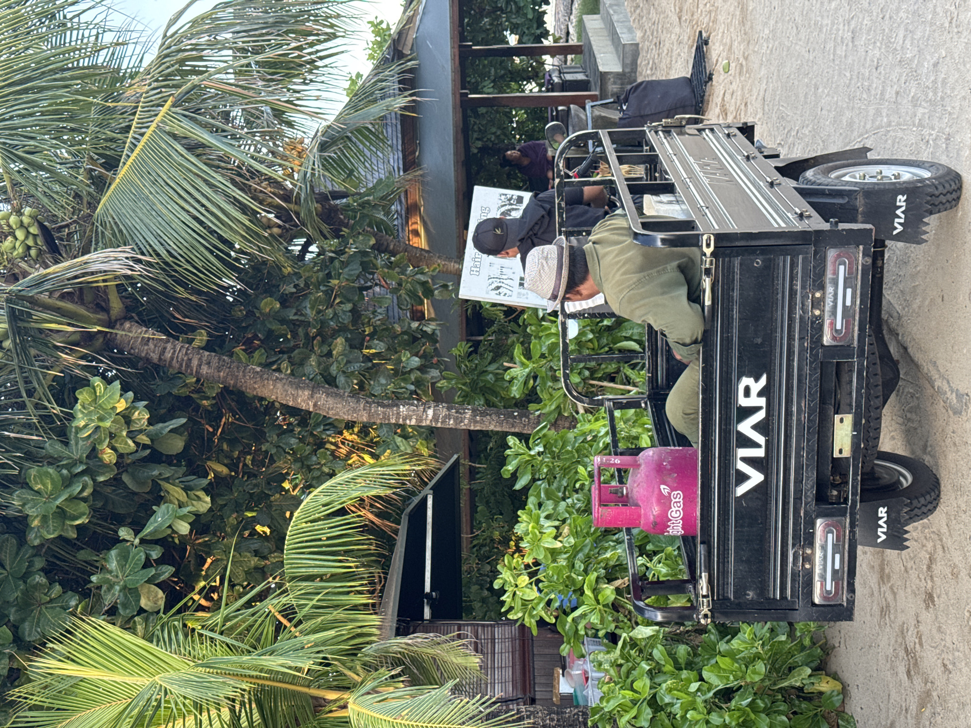 This image captures a scene in an unknown location, featuring a three-wheeled vehicle parked partially on a sandy or concrete surface near a lush, tropical area. The vehicle, labeled "VIAR", contains two individuals: one in an olive-green shirt and light-colored hat, and another in a black shirt and baseball cap, reading a sign. A pink gas tank is attached to the back of the vehicle. Tropical vegetation, including palm trees with coconuts, surrounds the scene, partially obscuring the view of a building or structure in the background, where a third individual is present.