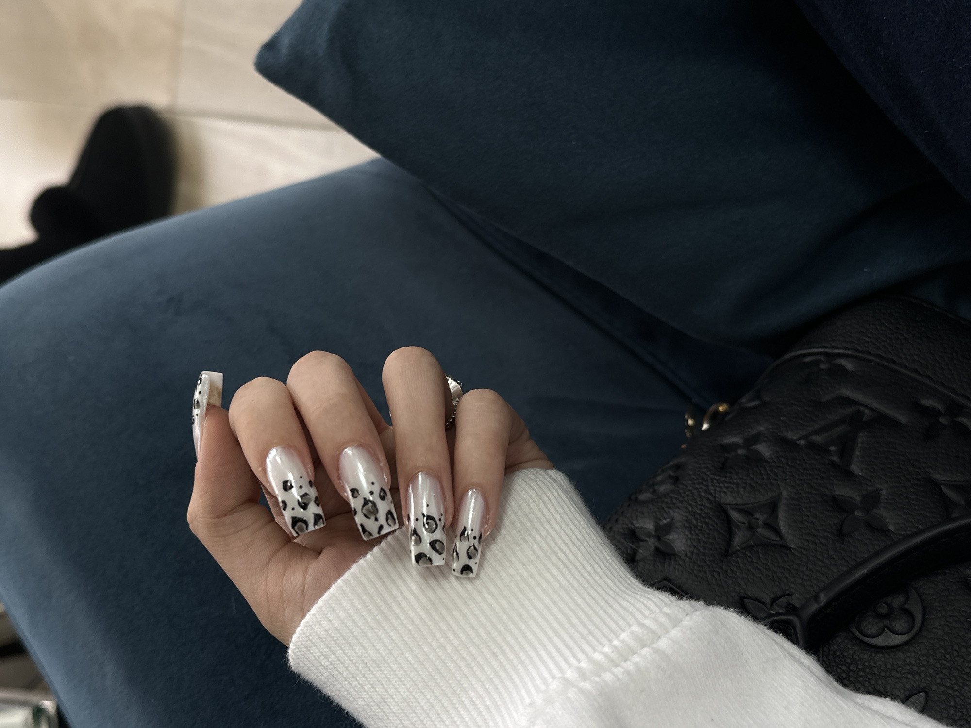 A person wearing a white sweater and sitting on a dark blue fabric-covered seat or cushion is displaying their hand with long, square-shaped nails featuring a white base with black leopard-print style spots. Adjacent to the hand is a black leather handbag with embossed logos. Behind the seat and hand, a dark-colored shoe is visible, and a partially visible floor is present. The location is not able to be determined based on this image.