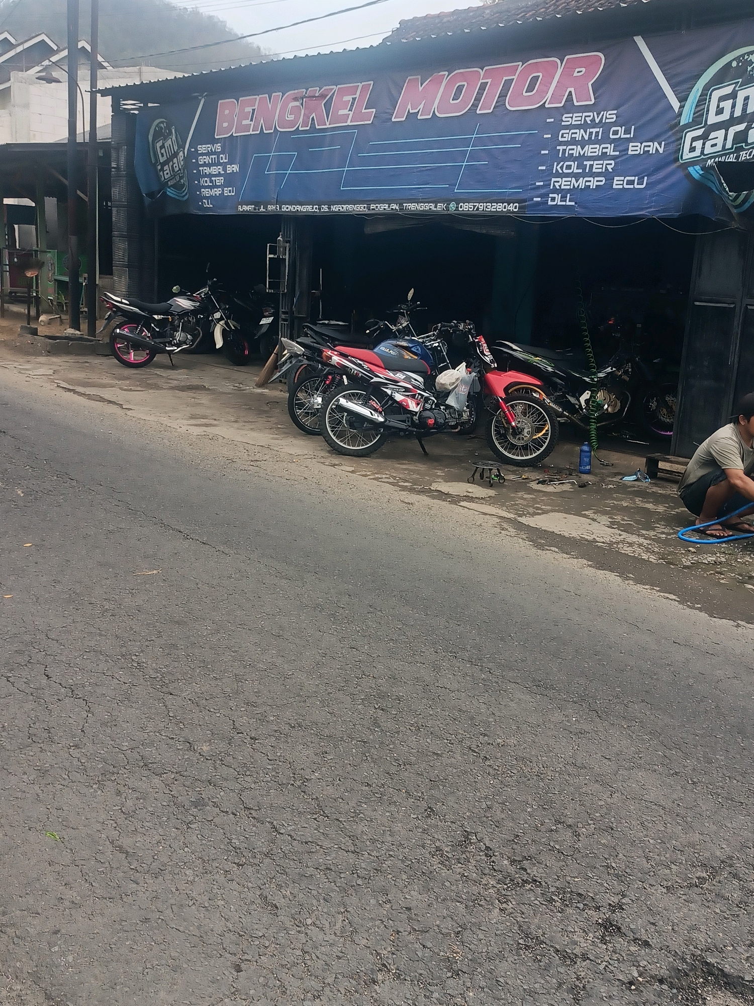 The image shows a motorcycle repair shop in Trenggalek, Indonesia. The shop's sign reads "BENGKEL MOTOR" and lists services such as oil changes, tire repair, and engine remapping. Several motorcycles are parked outside the shop, and a person is seen kneeling by the entrance, possibly working on a motorcycle. The shop appears to be located on a street in a rural or semi-urban area, with a damaged asphalt road in the foreground. The overall scene depicts a typical roadside motorcycle repair business.
