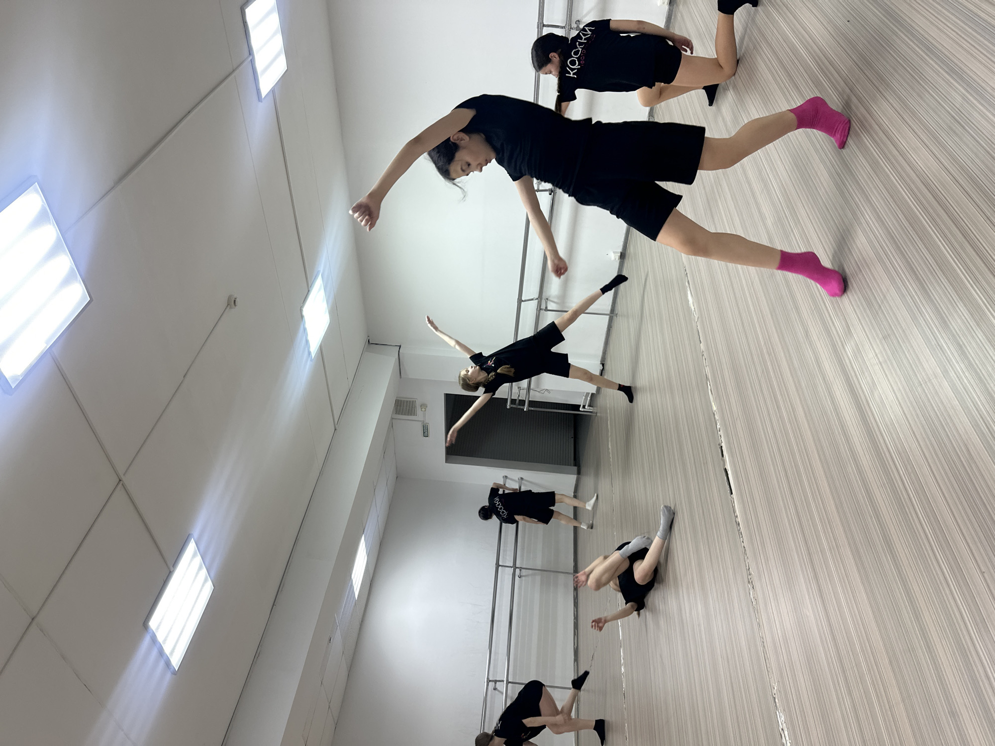 The image depicts a group of young people, likely dancers, engaged in a training session within a brightly lit indoor studio. The photograph is oriented diagonally, presenting the light-colored wooden or laminate floor as if it were a wall or ceiling, with the subjects positioned upon it.
There are at least five individuals, predominantly young females, dressed uniformly in black activewear consisting of t-shirts and shorts. They are captured in various dynamic and stretching poses, indicative of a dance or movement class. In the foreground, a person with dark hair is mid-pose, leaning back with arms extended and wearing bright pink socks. Another individual kneels behind them, while a third is seen with arms and one leg extended horizontally. Two more individuals are visible on the floor, one with legs raised and another further down the frame.
The studio features plain white walls and a white ceiling equipped with several rectangular recessed fluorescent light fixtures, which cast strong illumination and visible light rays. A ballet barre runs along a white wall in the background, confirming the setting as a dance or exercise facility. On the back of one person's black t-shirt, the Cyrillic text "КРОССКИ" is clearly visible, translating to "Sneakers" or "KROSSKI." The atmosphere suggests focused activity and practice. The time of day is not evident due to the artificial lighting. This scene takes place in Votkinsk, Russia.