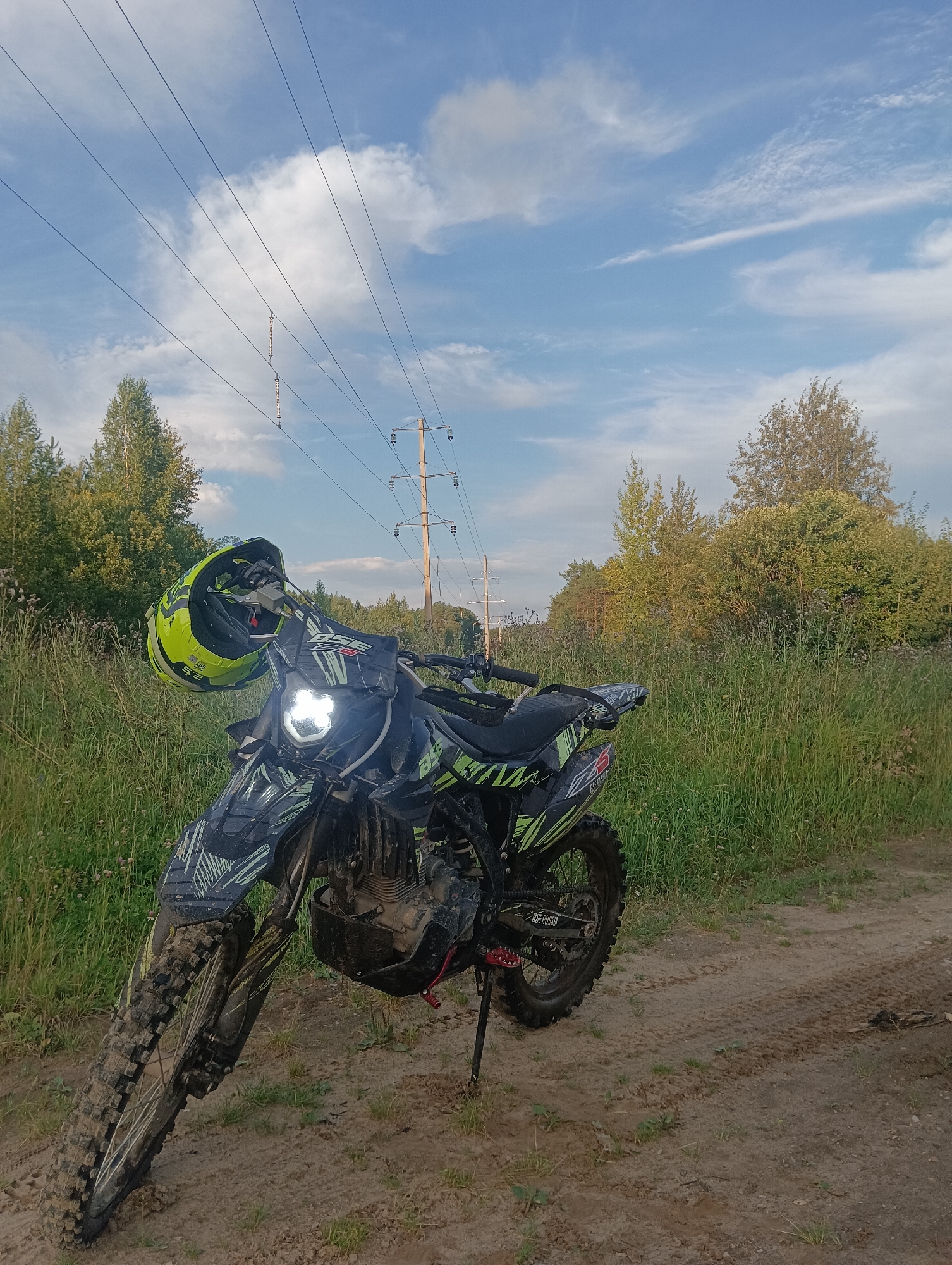 An outdoor, medium-shot, eye-level, color photograph shows a dirt bike parked on a dirt path with a field of tall grass to the left and a line of trees in the background. A yellow and black helmet is hanging from the handlebars of the dirt bike. The sky is blue with scattered white clouds and power lines stretch across the upper portion of the image. The dirt bike is black with green accents and appears to have been recently ridden through mud. The headlights of the dirt bike are illuminated. The ground is a mixture of dirt and grass. The overall scene appears to be in a rural or natural setting, possibly a trail or unpaved road.
