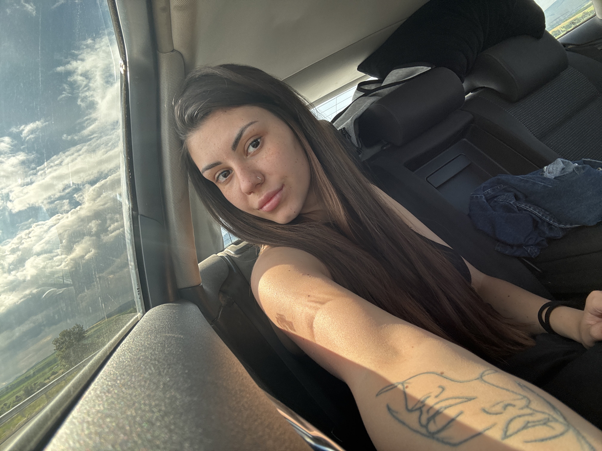 A young woman with long, dark brown hair and a nose piercing takes a selfie inside a car, looking directly at the camera with a calm expression. She has visible freckles on her face. Strong, warm sunlight streams into the car from the left, illuminating her face and casting a distinct shadow across her arm. On her left forearm, a line art tattoo depicting a face is visible. She wears a dark-colored top, and a black hair tie is on her right wrist.
The car's interior features dark seats with headrests, and a crumpled denim garment rests on the back seat. Through the car window on the left, a bright, cloudy sky with large white clouds dominates the view. Below the clouds, a green landscape with fields, scattered trees, and a visible road or highway stretches into the distance, suggesting the car is traveling through a rural area. The scene depicts a sunny day, likely late afternoon or early evening, in Hrabarsko, Bulgaria. No visible text is present in the image.