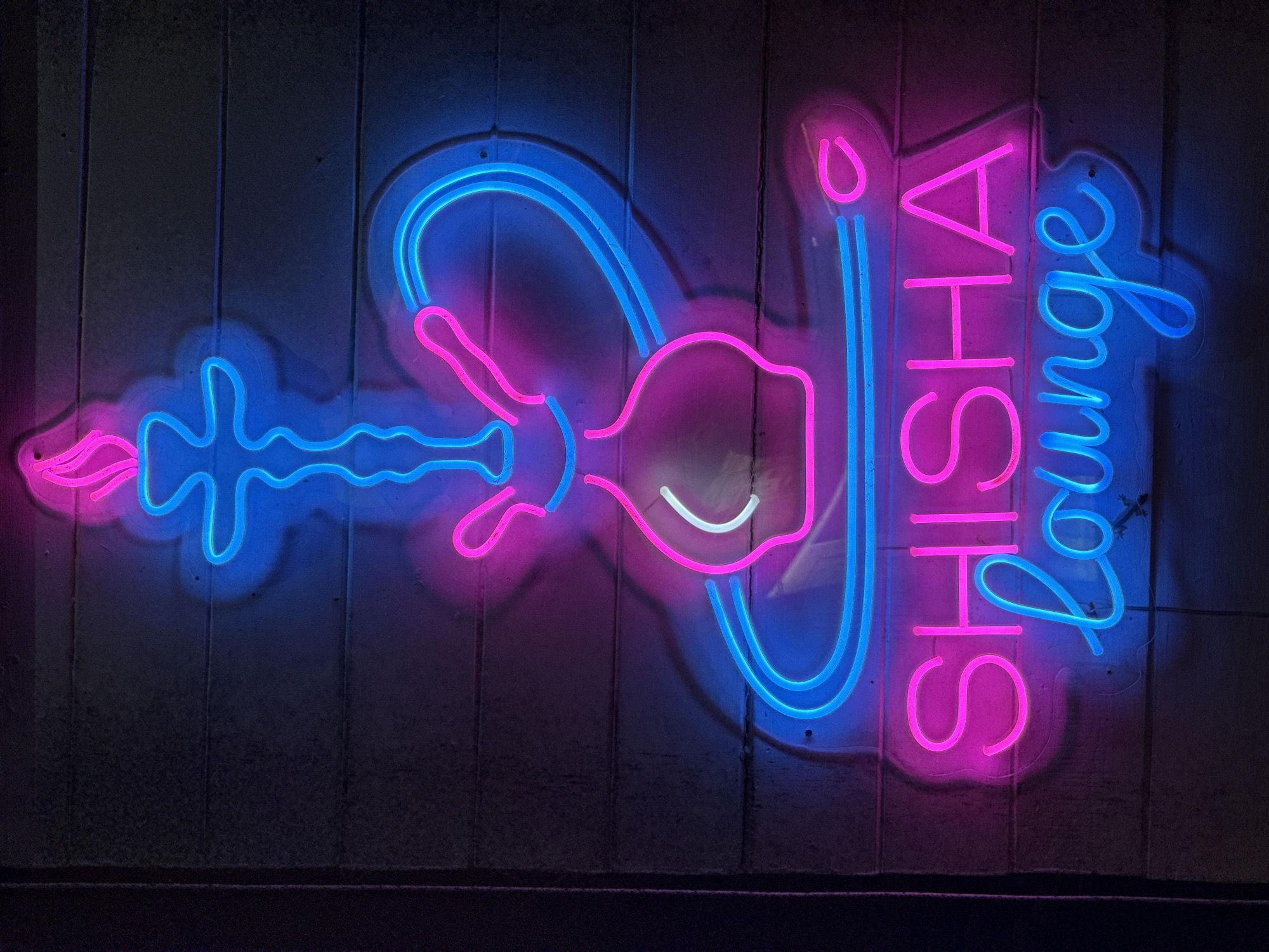 A neon sign displays a hookah and the words "SHISHA lounge" in pink and blue neon lights. The sign is mounted on a textured, dark gray wall. The hookah graphic is depicted in blue neon, with pink accents representing smoke. The text "SHISHA" is in bright pink neon, and "lounge" is in bright blue neon in a cursive font. The wall appears to be made of horizontal planks. This sign is likely located in Baan Bo Phut, Thailand, indicating a place where shisha is served.
