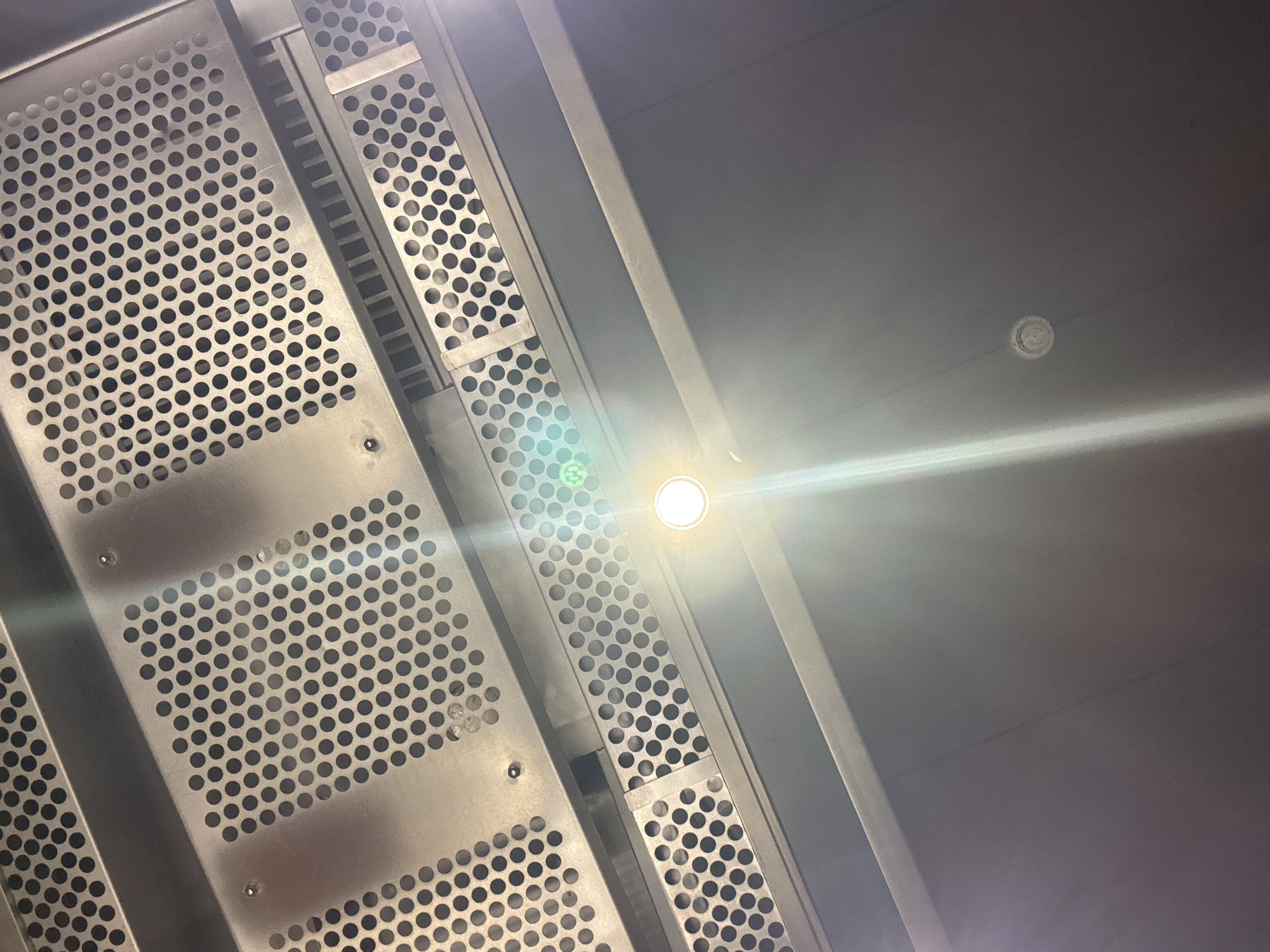 The image is a low-angle shot of a metal, perforated structure, likely part of a ventilation system or ceiling element. The structure has a grid-like pattern of circular holes, with some sections appearing to be staggered. The metal has a matte finish and reflects ambient light. The background is a plain, greyish surface, probably a ceiling. A bright light source, likely an LED, is visible in the center of the image, with a noticeable lens flare. The location is unknown and cannot be determined from the image. No people, subjects, objects, actions, or interactions are visible in the image other than the structural elements and the light source.