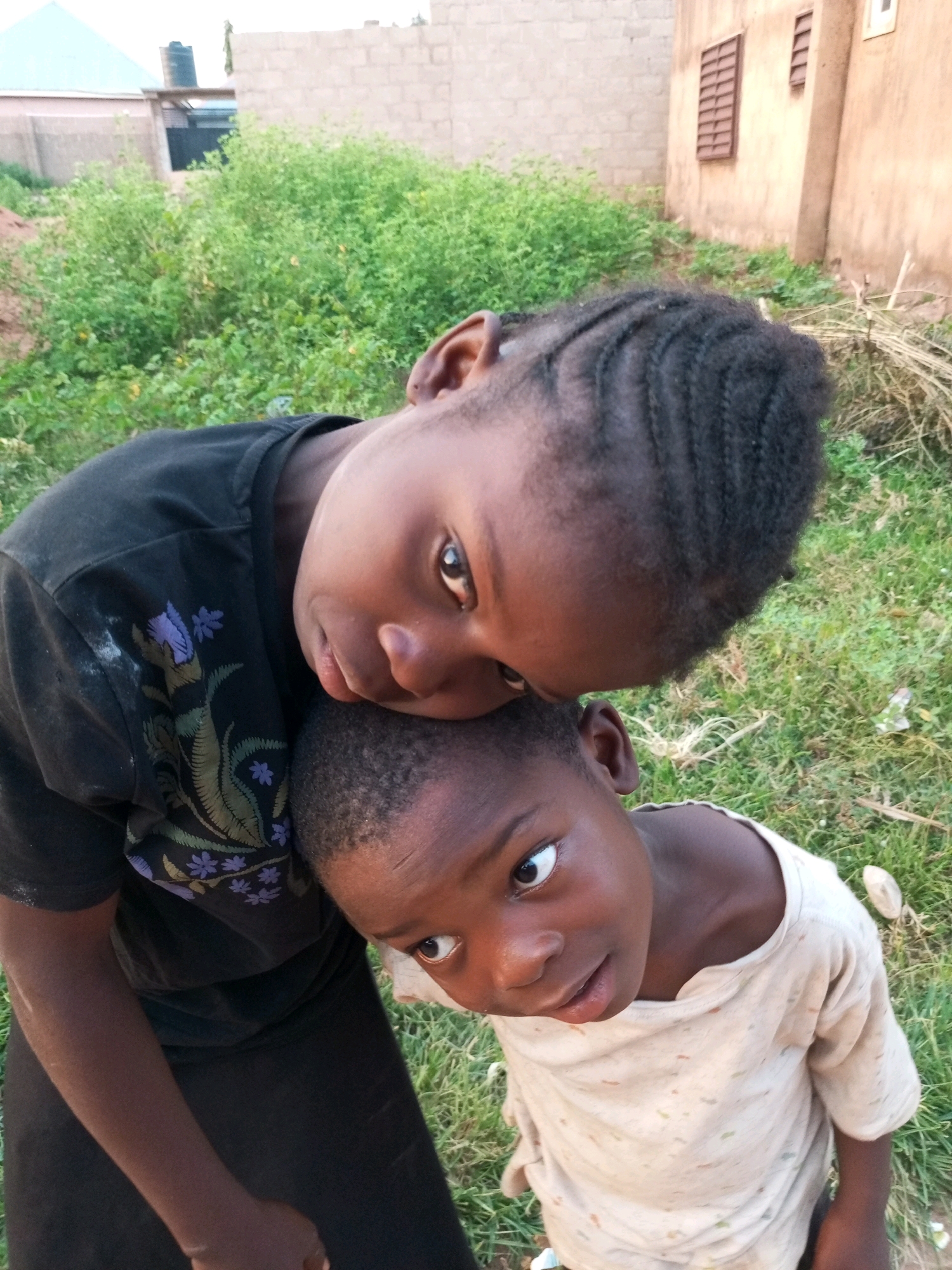 In an outdoor setting, possibly within a residential area in Tempure, Nigeria, two young children are the central subjects of the photograph. The child in the foreground, wearing a light-colored t-shirt, is looking up and to the left with wide eyes. Their facial features are clearly visible, with dark hair and skin. The second child, whose head is resting on the first child's shoulder, is looking towards the camera. This child is wearing a black t-shirt with a floral pattern. Their face is partially obscured but their eyes are visible. The background consists of green grass and some dry vegetation, with a textured building wall and a window visible in the upper right quadrant, suggesting an urban or semi-urban environment. The lighting suggests it is daytime.