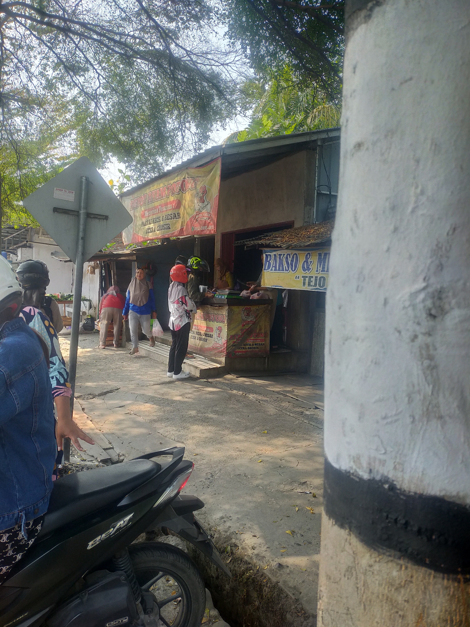 The image depicts a street scene in Slawi, Indonesia, likely in front of a food stall. In the foreground, a black motorcycle is partially visible on the left side of the frame, with its rear wheel and part of the seat showing. A person wearing a denim jacket and patterned pants is standing next to the motorcycle, with their hand resting on the seat.
The midground of the image features a small building that appears to be a food stall. A yellow banner with red text hangs across the front, advertising a food business. Several people are present at the stall. One person in a red helmet and white shirt is standing at the counter, while another person in a blue shirt and grey pants walks away from the stall carrying a plastic bag. Two other individuals, one in a red top and headscarf and another in a grey headscarf, are also visible near the stall. A smaller banner below the yellow one also displays advertising text. A signpost is visible on the left side of the street.
The background shows trees and foliage, suggesting an outdoor environment. The ground is paved and appears to be a street or sidewalk. The overall impression is a busy street corner with a local food vendor operating.