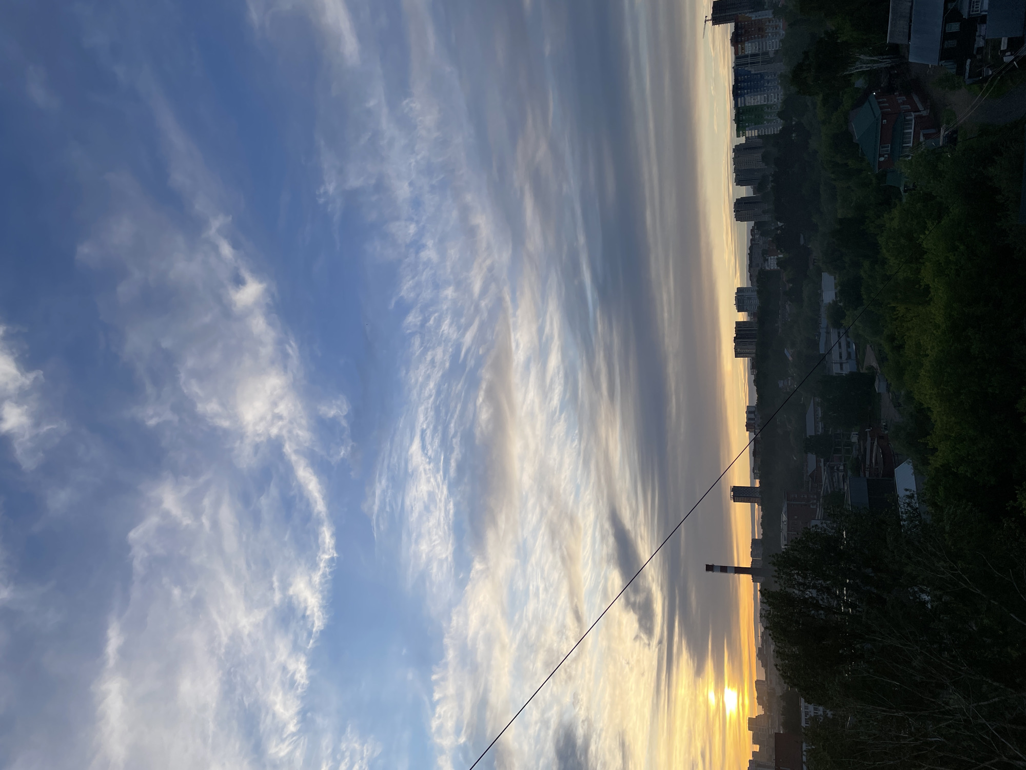 The image depicts a cityscape at sunset. The sky is filled with long, streaky clouds with shades of white, grey, and blue. The sun is setting on the horizon, casting a warm golden light across the city.
Below the horizon, there are buildings and trees. A tall smokestack is visible, a symbol of the industrial sector. Power lines are crossing the view.
The location is Izhevsk, Russia, based on the smokestack structure and cityscape.