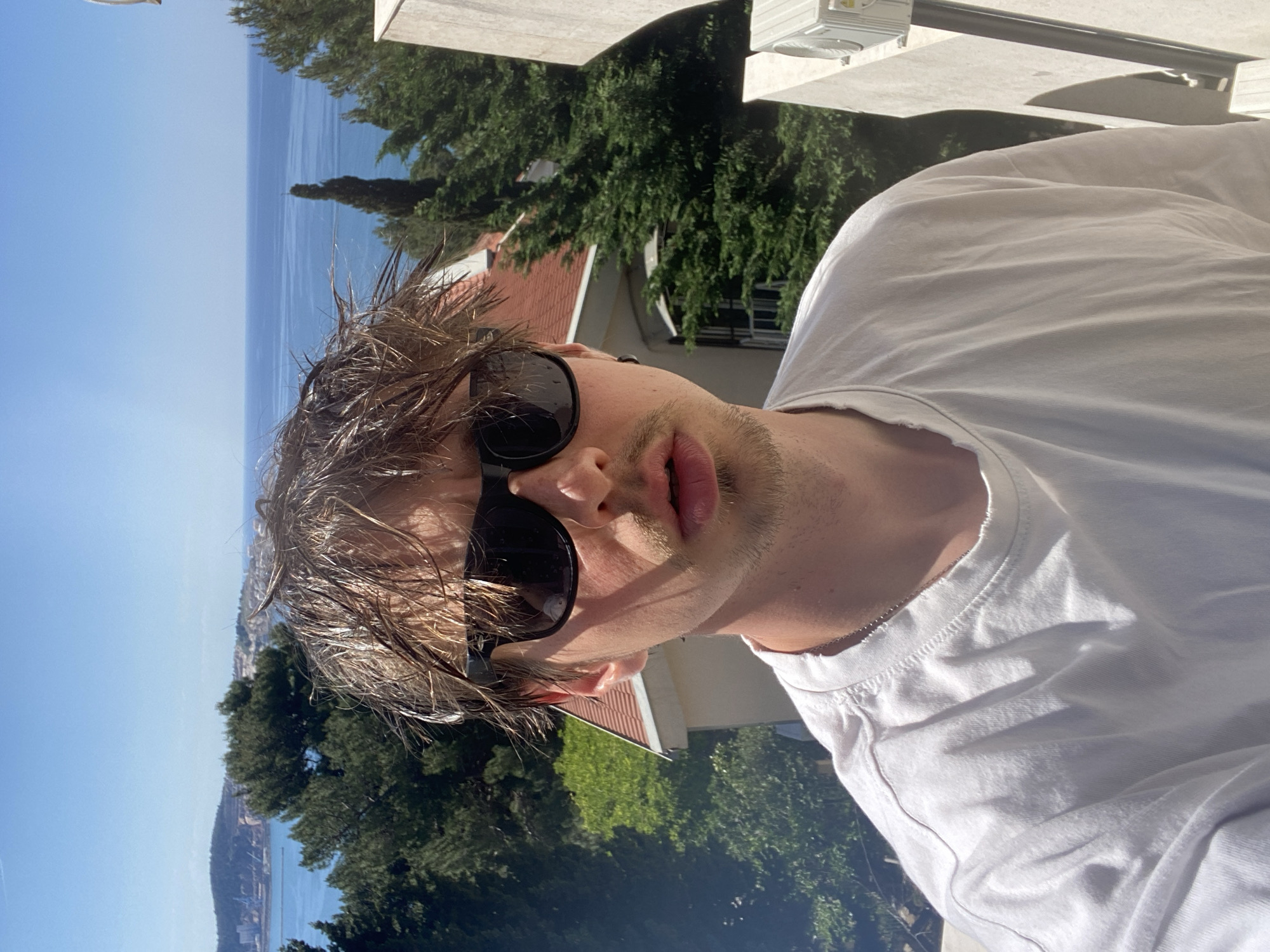 A young man is prominently featured in a bright, sunlit, diagonally oriented portrait, likely a selfie, against a scenic coastal backdrop. He has messy, light brown hair and is wearing dark sunglasses that obscure his eyes. A light stubble covers his chin and upper lip, and his lips are slightly parted. He wears a plain white t-shirt, with a thin silver chain faintly visible around his neck.
The background displays a serene coastal landscape under a clear, bright blue sky. A vast expanse of calm, blue sea stretches across the mid-ground, reflecting the strong sunlight. Lush green trees, including tall, slender conifers and other dense foliage, dot the hillsides, interspersed with buildings featuring distinctive red-tiled roofs. These architectural elements and natural features are characteristic of the Adriatic coast, specifically in Šušanj, Montenegro. A distant landmass or mountain range is visible across the bay. The strong lighting and clear sky indicate a bright, sunny day, likely midday or early afternoon. No visible text is present in the image.