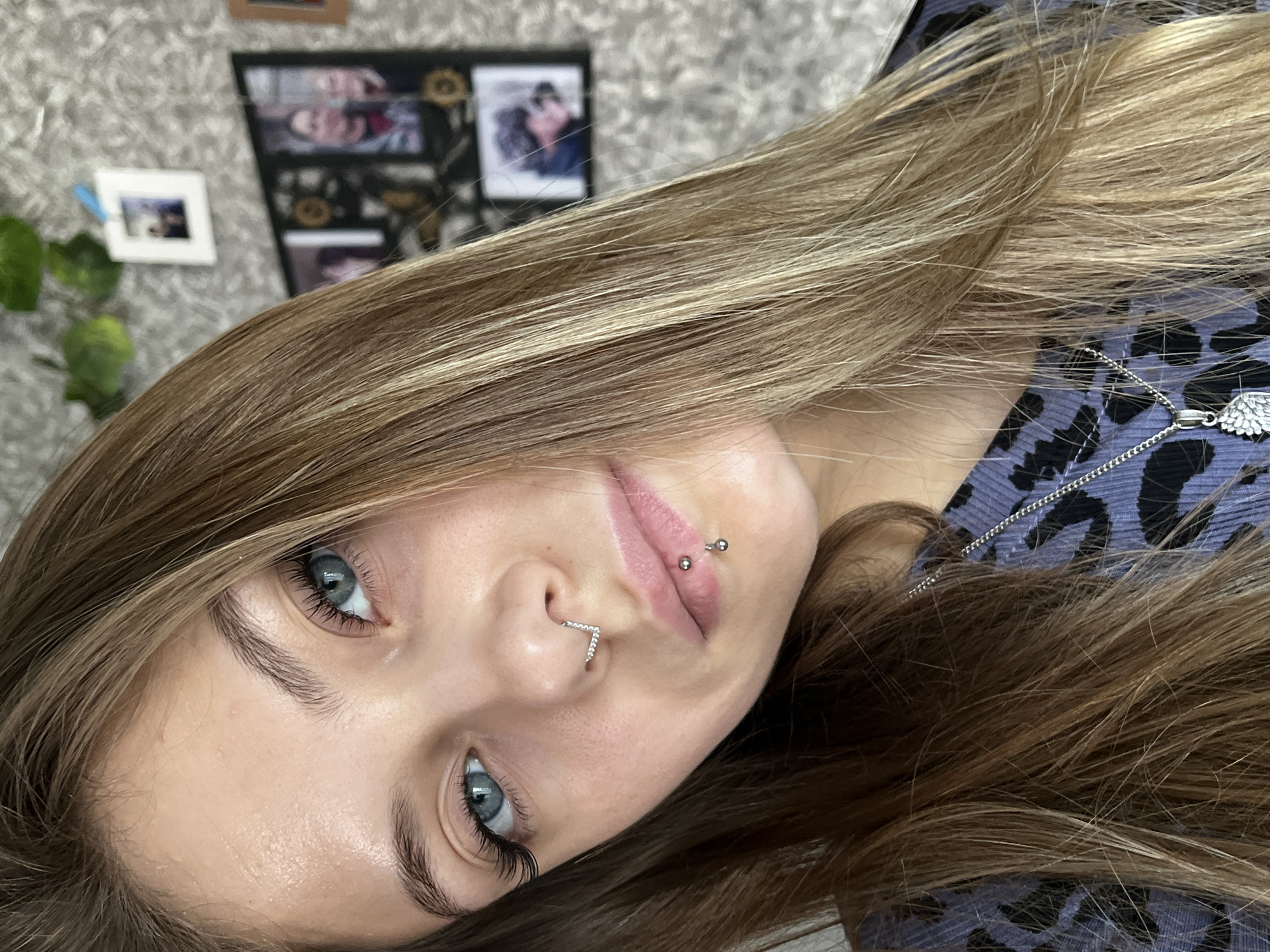 A close-up portrait of a young woman with light brown hair and blue eyes. She has multiple piercings, including a septum piercing and lip piercings. She is wearing a patterned shirt and a necklace with a wing pendant. The background is blurry, showing a wall with framed pictures and a houseplant. The image does not appear to be set in Фоки, Russia.