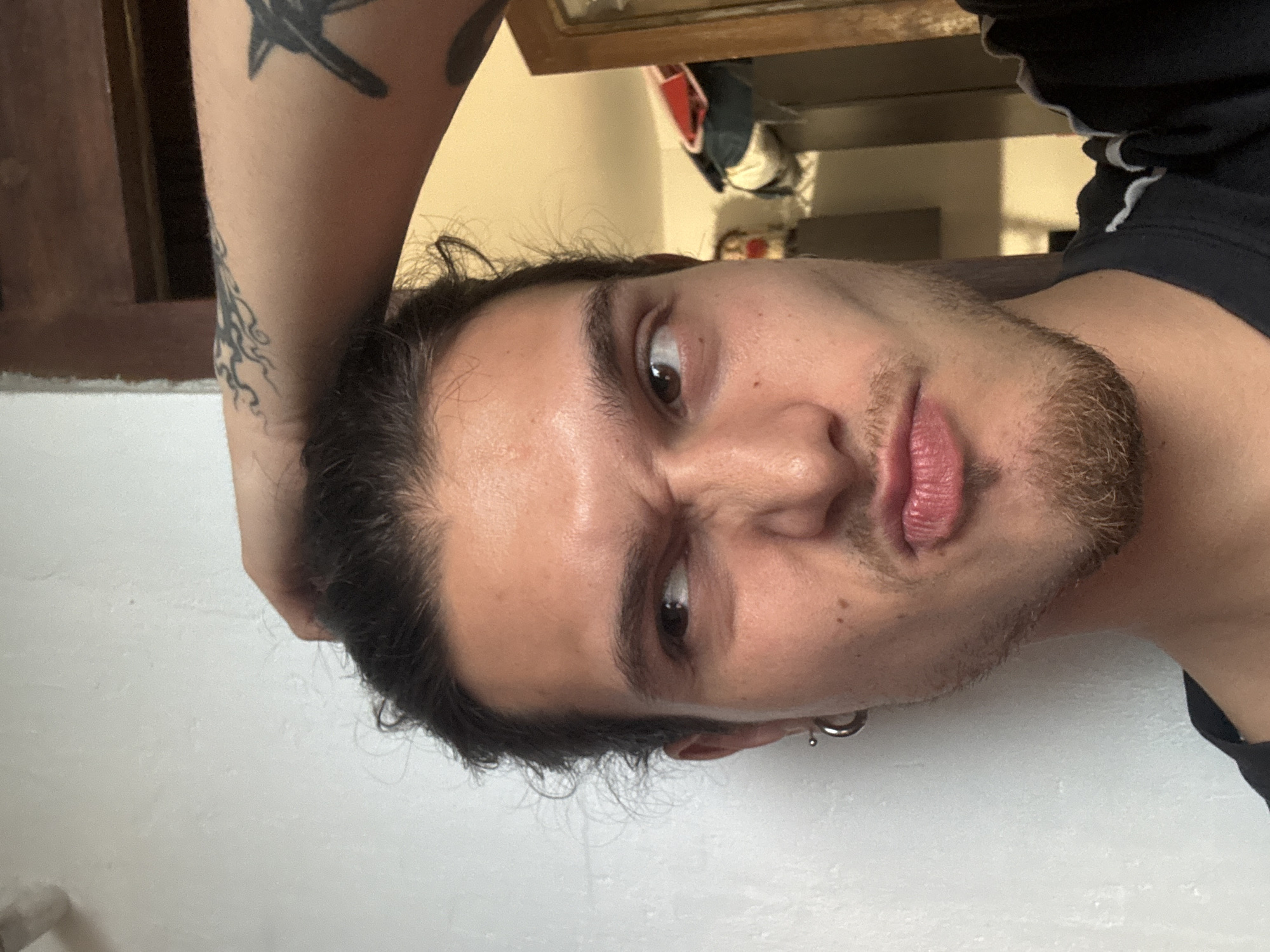 A man is posing for a selfie. He is indoors, with a view of an interior featuring a doorway, and a cabinet-like object in the background. The man has long dark hair, facial hair, and fair skin. He is wearing a black shirt and has tattoos on his left arm. The man appears to be holding his head up with his arm. The photograph's orientation is rotated, making the background appear vertically oriented. There are no details to suggest this photo was taken in Sofia, Bulgaria.