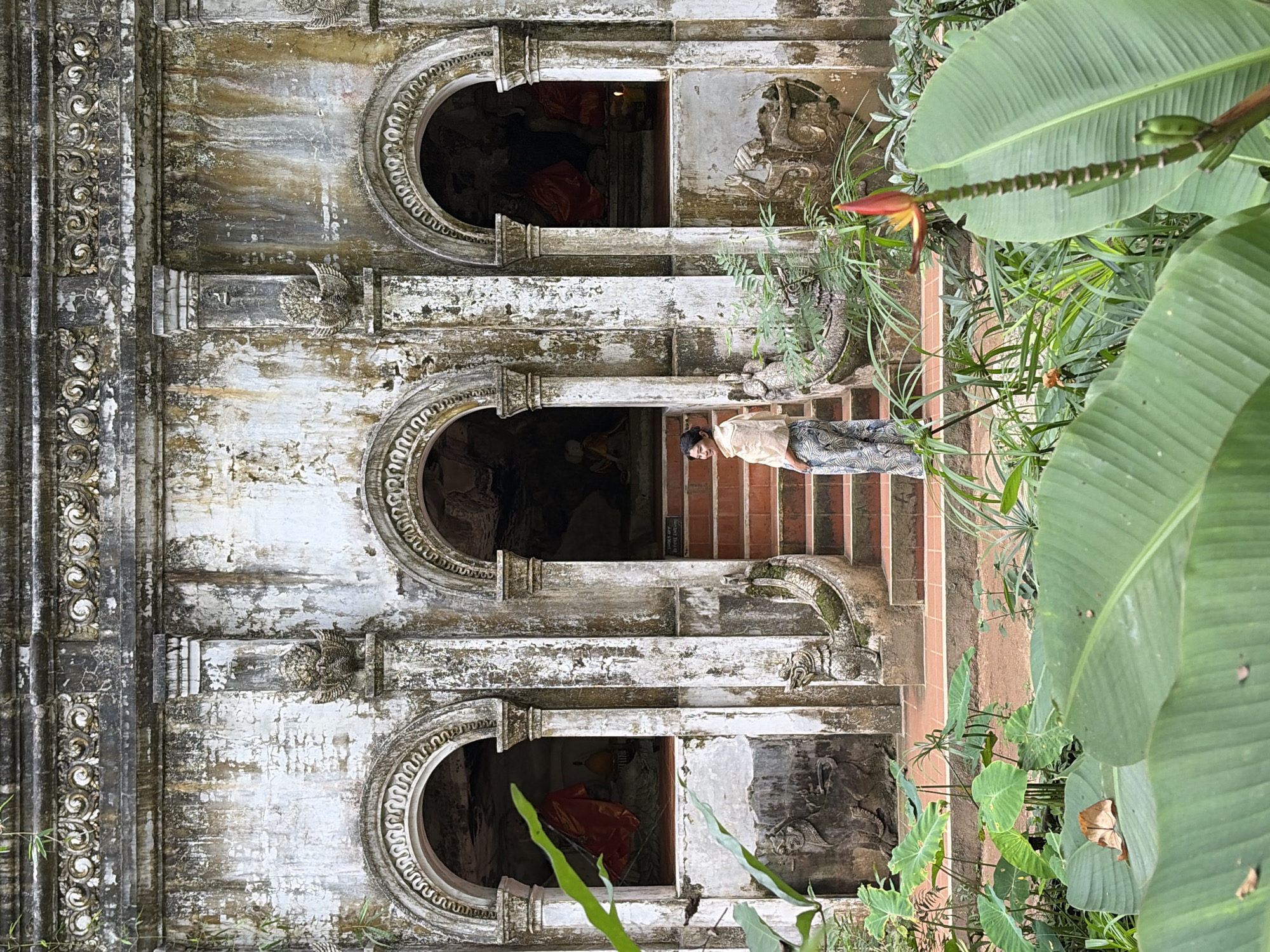 The image shows a woman standing in front of an old, weathered stone building with arched doorways. The building appears to be a temple or shrine, with statues or religious figures visible within the doorways. The woman is wearing a light-colored shawl and patterned pants. She is looking towards the left side of the frame. The building is adorned with intricate carvings and ornate details. The area in front of the building is lush with vegetation, including large tropical leaves and a flowering plant. The ground is covered in reddish-brown earth. The scene suggests an outdoor setting in a tropical climate. The architectural style and the presence of religious figures indicate a cultural or historical site, likely in Southeast Asia.