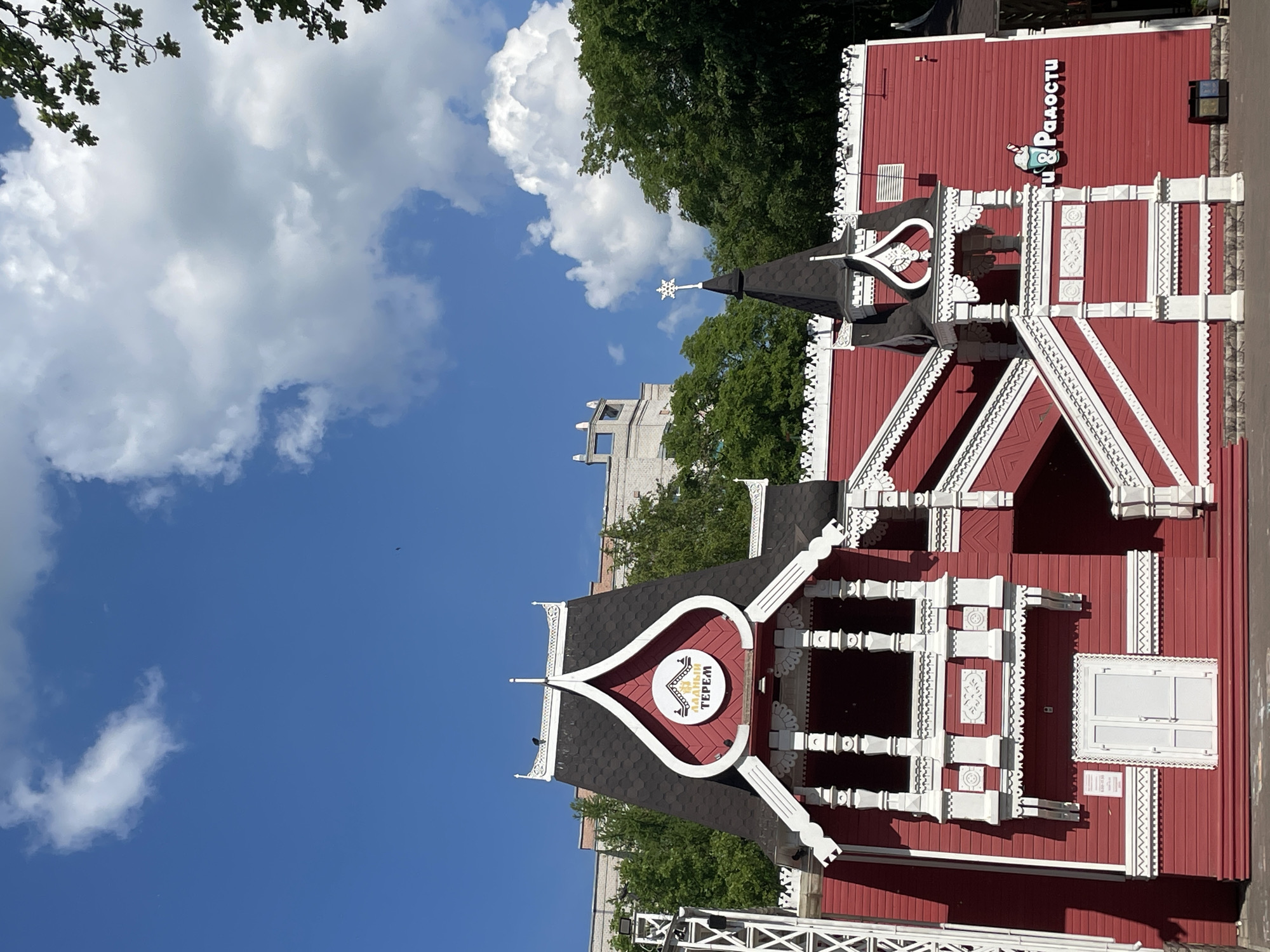The image captures a bright, sunny day in Vologda, Russia, with a vibrant red building featuring traditional Russian architectural elements. The building has a steeply pitched, dark roof with ornate white trim. A prominent sign on the front of the building reads "ЛАДНЫЙ ТЕРЕМ" in gold lettering within a circular emblem. To the right, another sign displays the words "ПРИЯТНОСТИ & РАДОСТИ" with a graphic of a milkshake or ice cream cone. White decorative railings and intricate wooden carvings adorn the exterior, particularly around the balconies and staircases. Green trees are visible behind the building, and a light-colored building with a modern architectural style stands in the background. The sky is a clear blue with scattered fluffy white clouds. The scene appears to be an outdoor area, possibly a park or a public space, with a white door leading into the red building. The overall impression is one of cheerful and culturally inspired design.